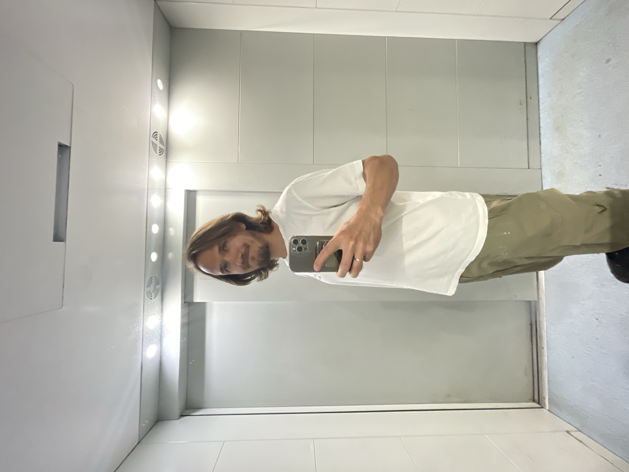 A medium shot captures a man with shoulder-length brown hair and a short beard taking a selfie in a modern elevator. He is wearing a white t-shirt and khaki pants, with a ring on his left ring finger. The elevator has white paneling on the walls and ceiling, with recessed lighting fixtures. A large, grey elevator door is visible behind him. The floor of the elevator is light grey and appears to be linoleum. The man is holding a silver smartphone in his right hand, which is angled to capture his reflection.