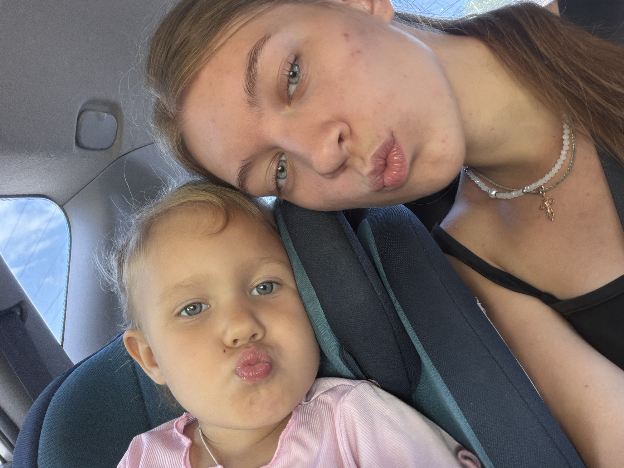 A young woman and a toddler are in the back seat of a car, both with their lips puckered for a kiss. The toddler, wearing a pink shirt, is nestled in a car seat to the left, while the woman, wearing a black tank top and a beaded necklace with a cross pendant, is on the right. The interior of the car is visible, with the ceiling and a rearview mirror in the background. The lighting suggests it is daytime. There are no specific landmarks indicating Rostov-on-Don, Russia.