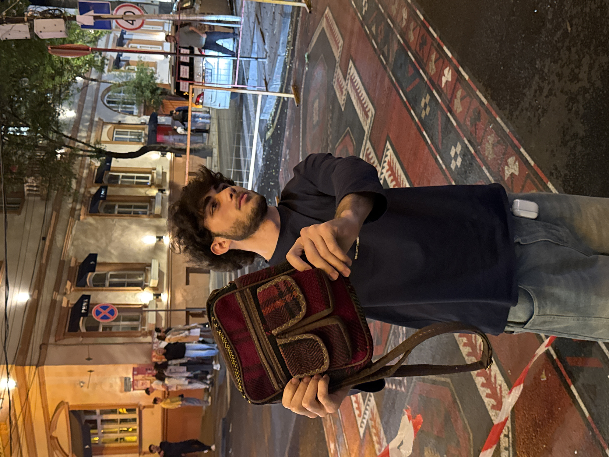 A person with dark hair and a beard is holding a reddish-brown bag with decorative stitching in their hands. The person is wearing a navy blue shirt and light blue jeans. An Apple brand earbud case is partially visible in the person’s pocket. The person is standing on a street decorated with a large, painted rug pattern. In the background, there are buildings with windows and signage. Other people are visible further in the background. There are streetlights providing illumination. The location is Yerevan, Armenia.