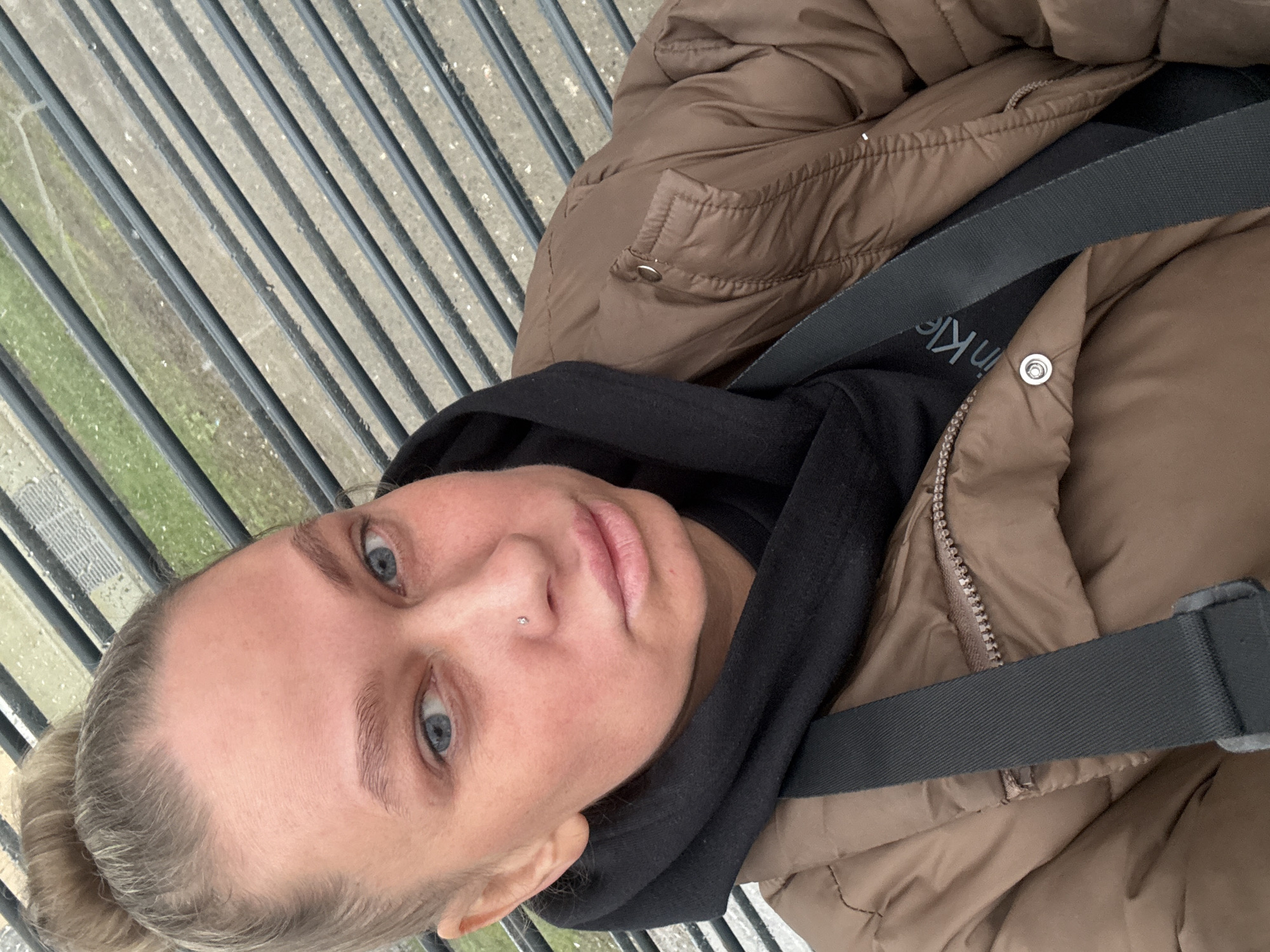 A woman with blonde hair in a bun, light blue eyes, and a nose piercing is taking a selfie. She is wearing a black hooded sweatshirt under a light brown puffer jacket. A black strap with white text crosses her torso. The background shows a metal railing overlooking a concrete area with some greenery visible. The location is likely the pedestrian bridge above stantsiya Perm I in the city of Perm, Russia.