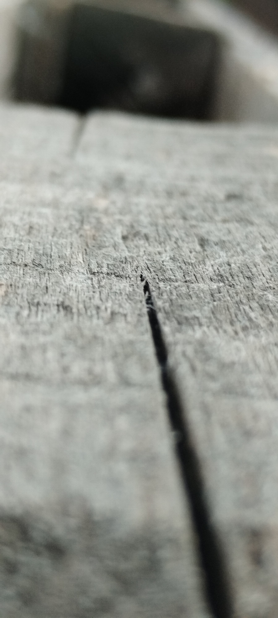 The image is a close-up, low-angle shot of a weathered, gray wooden surface, likely a plank or tabletop. A prominent, dark crack runs vertically through the center of the frame, widening slightly as it moves away from the viewer. The wood grain is visible, showing lines and textures consistent with age and exposure. In the background, out of focus, are dark, indistinct shapes that could be furniture or other objects. The lighting is dim, casting shadows within the crack. The scene appears to be outdoors or in an area with natural light, given the diffused quality of the illumination. No people or specific landmarks are visible. The setting is not identifiable as Yaroslavskaya, Russia, or any other specific location.