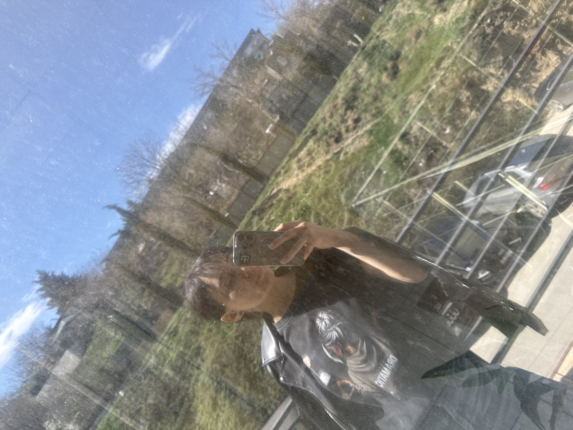 A person is taking a selfie in a glass window. The reflection shows a partial view of the person wearing a black leather jacket, and a t-shirt with a design. A phone is in their hand. The background reflection shows a landscape with buildings, trees, and vegetation. Reflected in the glass is a vehicle on a driveway or road. The location is Tbilisi, Georgia.
