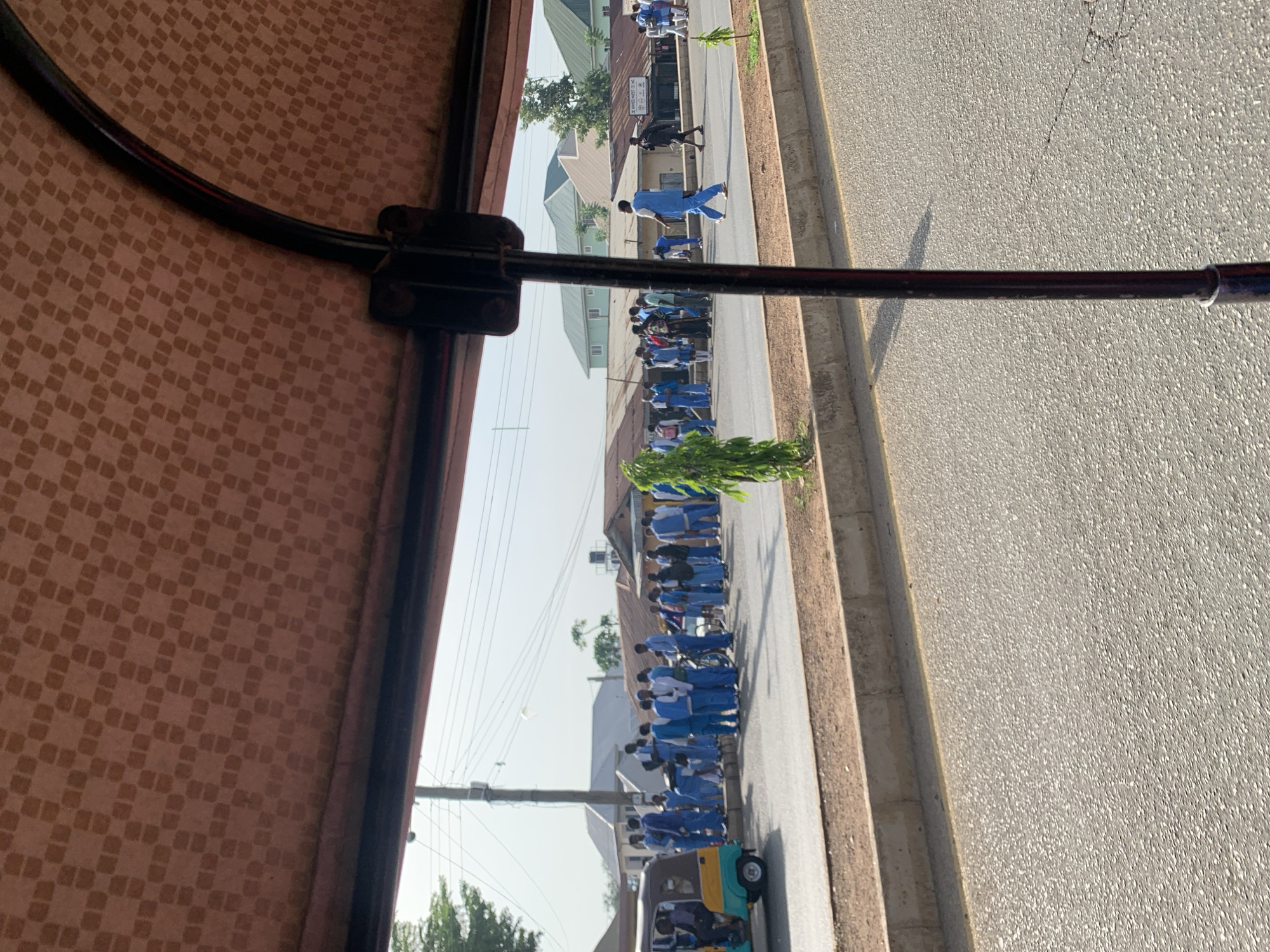 A line of people wearing blue and white uniforms march along a street in New Stadium, Nigeria. The photo is taken from inside a vehicle, possibly a bus or car, as indicated by the brown patterned interior and metal bar in the foreground. The street is paved and has a curb. Buildings line the street, and power lines are visible in the sky. A three-wheeled vehicle is at the end of the line of marchers.