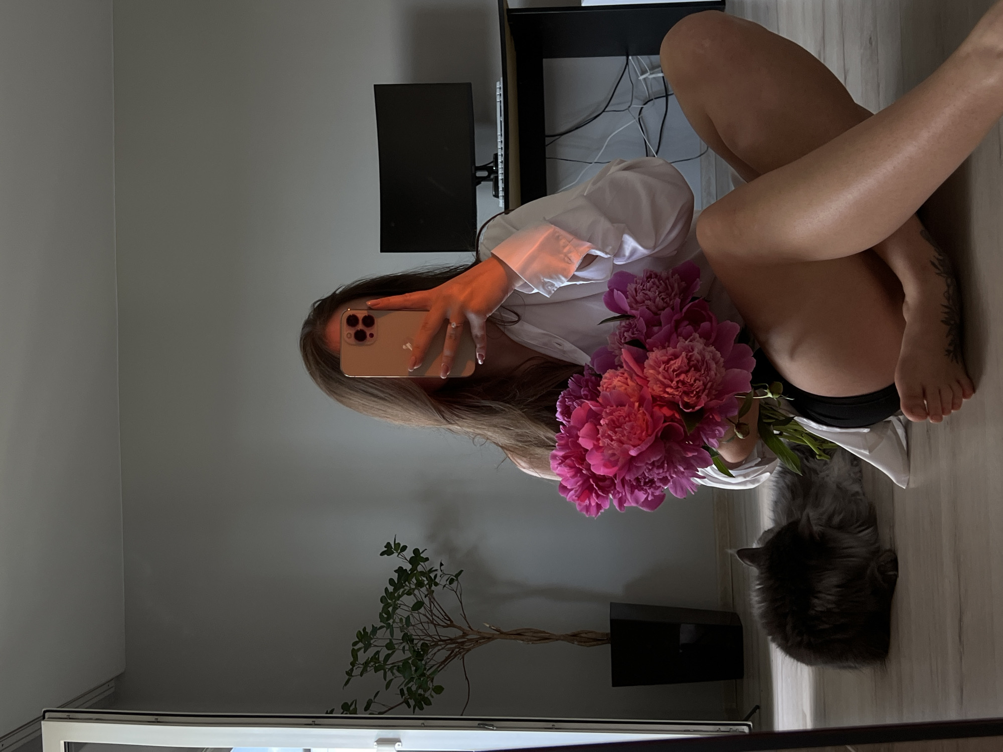 A young woman is taking a selfie with a bouquet of pink peonies. She is sitting on a wooden floor, wearing a white oversized shirt and black shorts. Her legs are crossed. To her left, a gray fluffy cat is grooming itself. In the background, there is a large plant in a black pot, a desk with a computer monitor, and a wall. The scene appears to be indoors, possibly in an apartment in Ternopil, Ukraine.