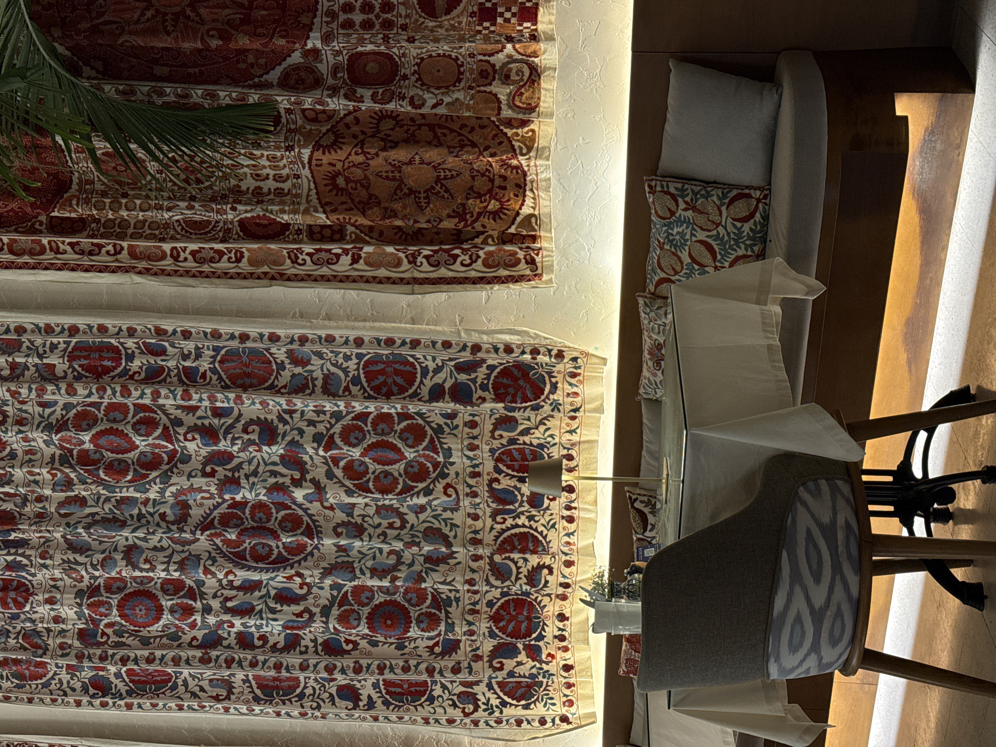 An indoor dining area, likely a restaurant or upscale lounge, is presented with a warm, inviting ambiance. The scene is devoid of people, suggesting a moment before or after service, or a quiet period.
On the left side of the image, two large, intricately patterned textiles, reminiscent of Central Asian suzani embroidery, are prominently displayed on a textured wall. The upper textile features rich red, orange, and brown hues with large circular floral motifs and paisley designs. Below it, a second textile exhibits vibrant red and blue floral and leaf patterns on a cream-colored background, also arranged in distinct circular and flowing designs. A verdant palm frond partially obscures the upper left portion of the top tapestry, introducing a touch of nature.
Separating the decorative wall from the dining area is a vertical strip of warm, recessed lighting that illuminates both the textiles and the textured wall. To the right, a dining setup includes a long, light-grey upholstered banquette adorned with cushions, one of which has a distinct red and blue pomegranate or floral pattern. A table, possibly with a glass top, is covered by a crisp white tablecloth. Several dining chairs are visible, including one with a grey upholstered seat and a patterned backrest featuring a light blue and grey ikat design. The legs and base of another chair, possibly with a black metal frame, are seen further right. A small potted plant adds a touch of greenery to the table setting.
The overall lighting is dim and artificial, casting a warm glow across the scene, particularly highlighting the architectural elements and the floor on the right, suggesting an evening or nighttime setting. The aesthetic, with its rich textiles and warm lighting, evokes a sense of cultural depth and sophisticated comfort, aligning with the location context of Dubai, United Arab Emirates. There is no clearly discernible text.