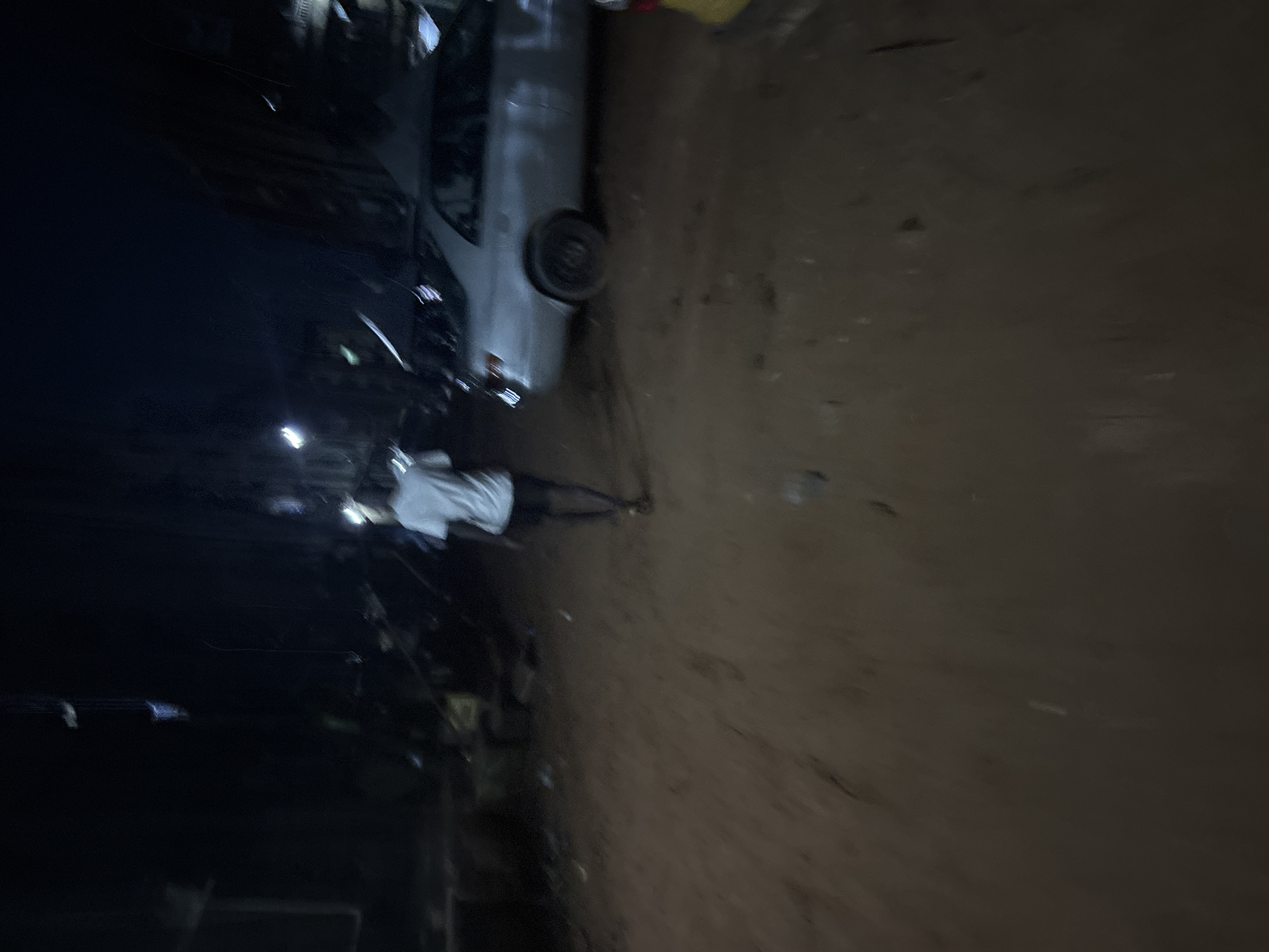 The image depicts a dimly lit street scene in Onitsha, Nigeria, at night. A single person, wearing a white t-shirt and dark shorts, walks away from the camera towards the right side of the frame. To the right of the person, a silver sedan is parked on the unpaved, muddy ground. The surroundings are dark, with some indistinct shapes of buildings or structures visible in the background, illuminated by what appear to be streetlights. The ground is uneven and appears to be made of red earth, with some debris scattered around. The overall impression is that of a rural or underdeveloped urban area at night, possibly experiencing low light conditions due to power outages or the time of day.