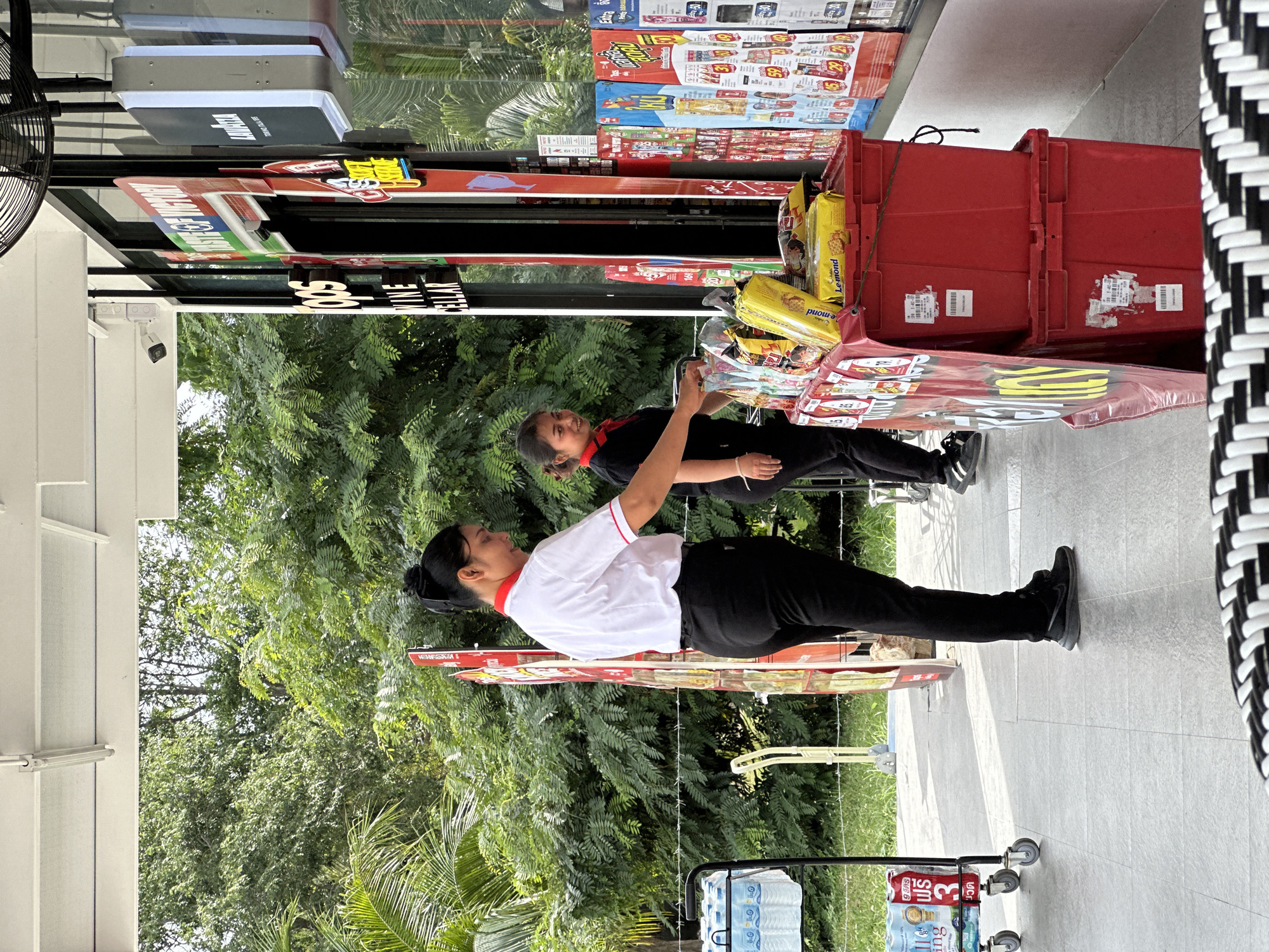 At an outdoor market stall in Koh Phangan, Thailand, two female vendors are assisting customers. One vendor, wearing a white shirt with red trim and black pants, stands near a cart laden with bottled water. The other vendor, in a black shirt with red trim and black pants, is interacting with a customer in front of a display of snacks and drinks. The stall features various advertising posters and bins filled with merchandise. The background is characterized by lush green trees and foliage.