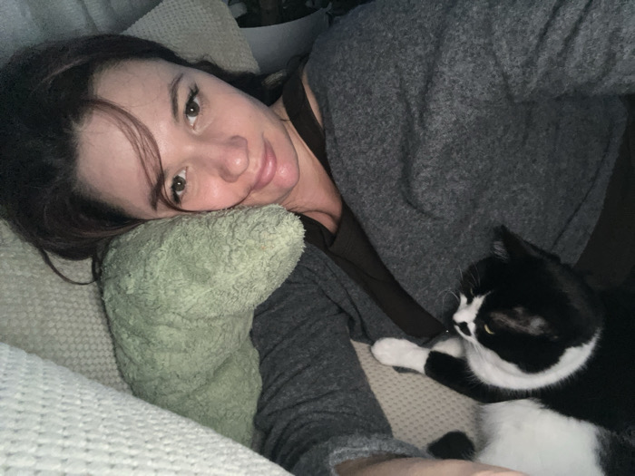 The image displays an adult female individual and a domestic cat within an interior setting. The female, possessing dark hair, is positioned on her left side, resting her head on a light green, textured pillow. She wears a dark grey long-sleeved top over a darker garment and gazes towards the camera with a subtle smile. A black and white tuxedo cat is situated to her right, resting on a light-colored, textured surface, likely a sofa or bed. The cat's head is oriented away from the camera, facing towards the female subject's left. The background is indistinct, confirming an indoor environment. The specific location within Budva, Montenegro, is not discernible from the provided visual information.