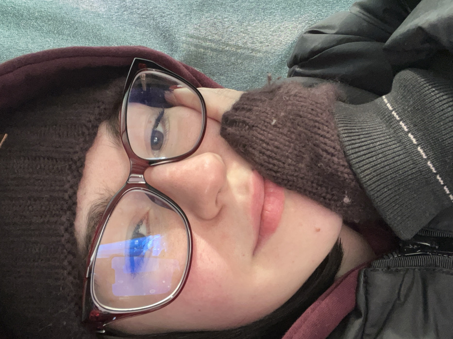 A close-up, eye-level portrait shows a single young female subject, angled slightly to the viewer's right. The subject wears dark maroon, possibly cat-eye style, eyeglasses; the left lens reflects distinct blue and white light sources. Her dark brown eyes are visible, with her gaze directed towards the viewer's left. Her light pink lips are slightly parted, forming a subtle smile. She has fair skin, with a light flush on her cheeks and small moles visible on her right cheek and chin. Her head is covered by a dark brown knitted beanie, and she wears a dark maroon hooded garment layered under a dark puffer jacket with a visible zipper. Her right hand, covered by a dark brown knitted fingerless mitt, rests against her chin and lower cheek. The background is an out-of-focus, textured light teal fabric or surface, suggesting an indoor setting. The specific city of Uman, Ukraine, cannot be determined from the visual information present in the image.