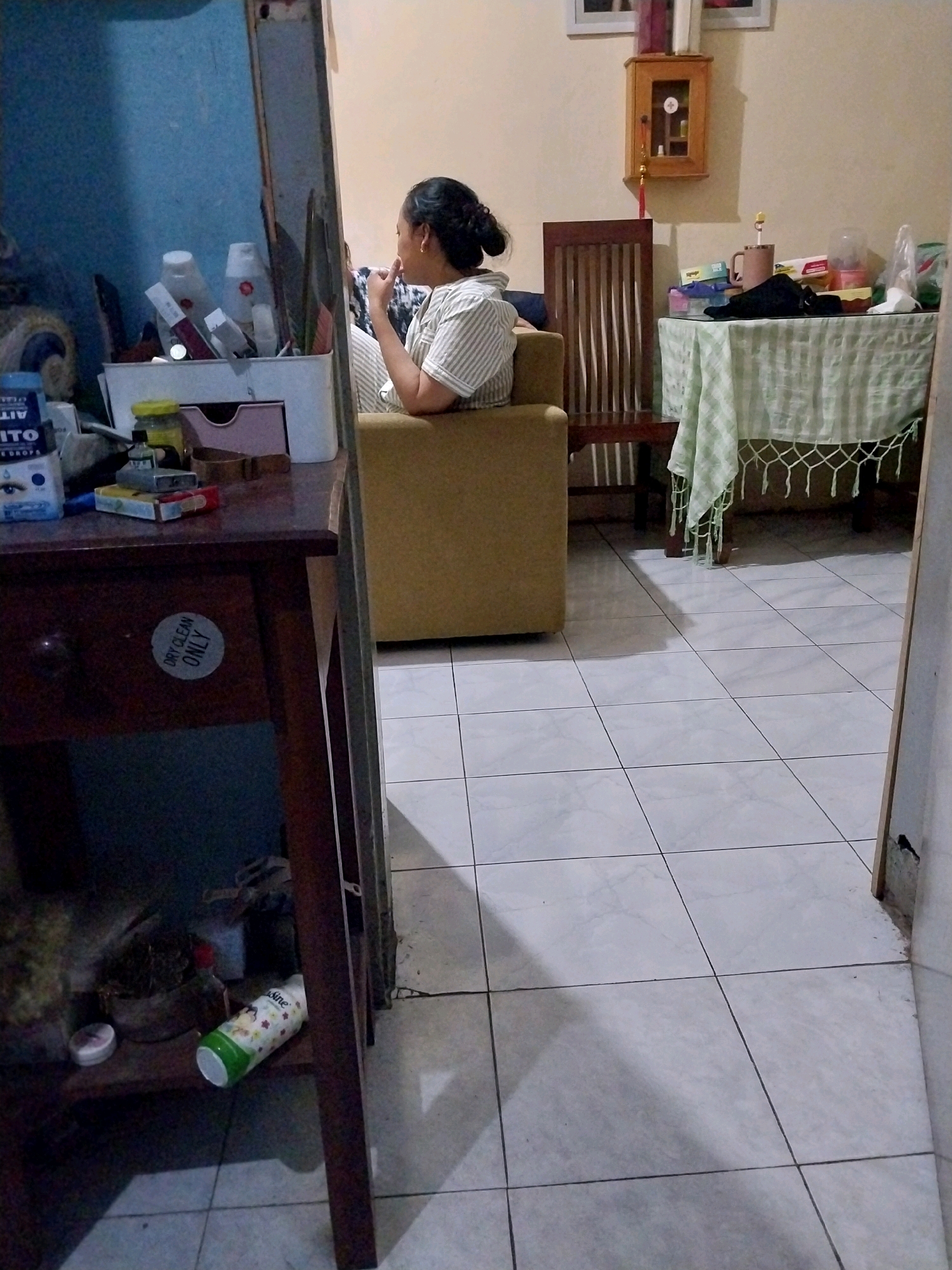 In a room with tiled flooring, a woman is seated on a mustard-colored armchair, facing away from the camera. She is wearing a striped shirt and her hair is pulled back in a bun. In front of the armchair, a dark wooden table stands, covered with a light green checkered tablecloth. On the table, several items are visible, including a mug, a black cloth, and some boxes. To the left of the armchair, a tall, dark wooden chair is positioned. The walls are painted a light beige color, and a small wooden cabinet with a glass door hangs on the wall above the table. To the left of the frame, a dark wooden table with drawers is visible, holding various bottles and containers, some of which appear to be cosmetic products. A round sticker with the words "DRY CLEAN ONLY" is affixed to one of the drawers. The scene appears to be an indoor domestic setting in the city of Ngadilangkung, Indonesia.
