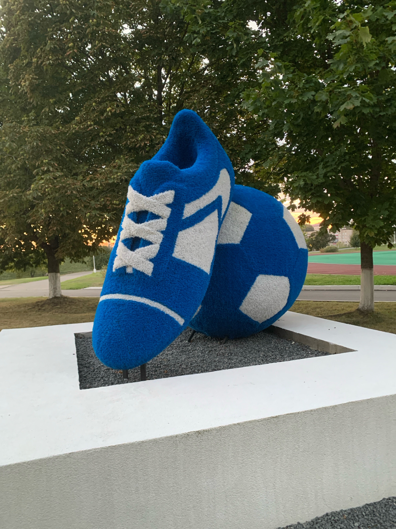 The image displays a large, stylized sculpture of a blue soccer shoe and a soccer ball, both covered in a fuzzy material. The shoe is in the foreground, positioned at an angle, with white laces and trim. The soccer ball, which is partially behind the shoe, is decorated with white hexagonal and pentagonal panels. Both objects are resting on a gravel bed within a white, rectangular, tiered platform. In the background, mature trees with lush green foliage are visible, with the warm glow of sunset filtering through their branches. Beyond the trees, a glimpse of a running track with red and green lanes can be seen, suggesting an outdoor sports facility. The overall setting appears to be a public space, possibly a park or an area adjacent to a stadium, within the city of Kapyĺ, Belarus. No people are present in the image, and the objects are static.