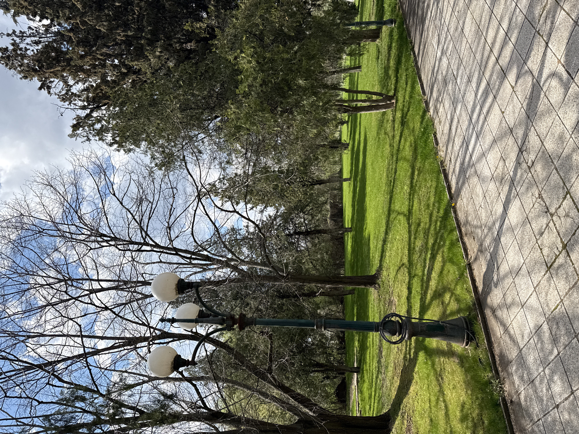The image depicts a park scene in Stara Zagora, Bulgaria. A three-globe lamp post stands prominently in the foreground against a backdrop of deciduous trees with bare branches reaching towards a sky with scattered clouds. Adjacent to the lamp post is a green lawn, and a paved walkway constructed of square blocks. The lighting suggests a sunny day, casting distinct shadows of the trees onto the grass and walkway. The overall scene suggests a public park setting.