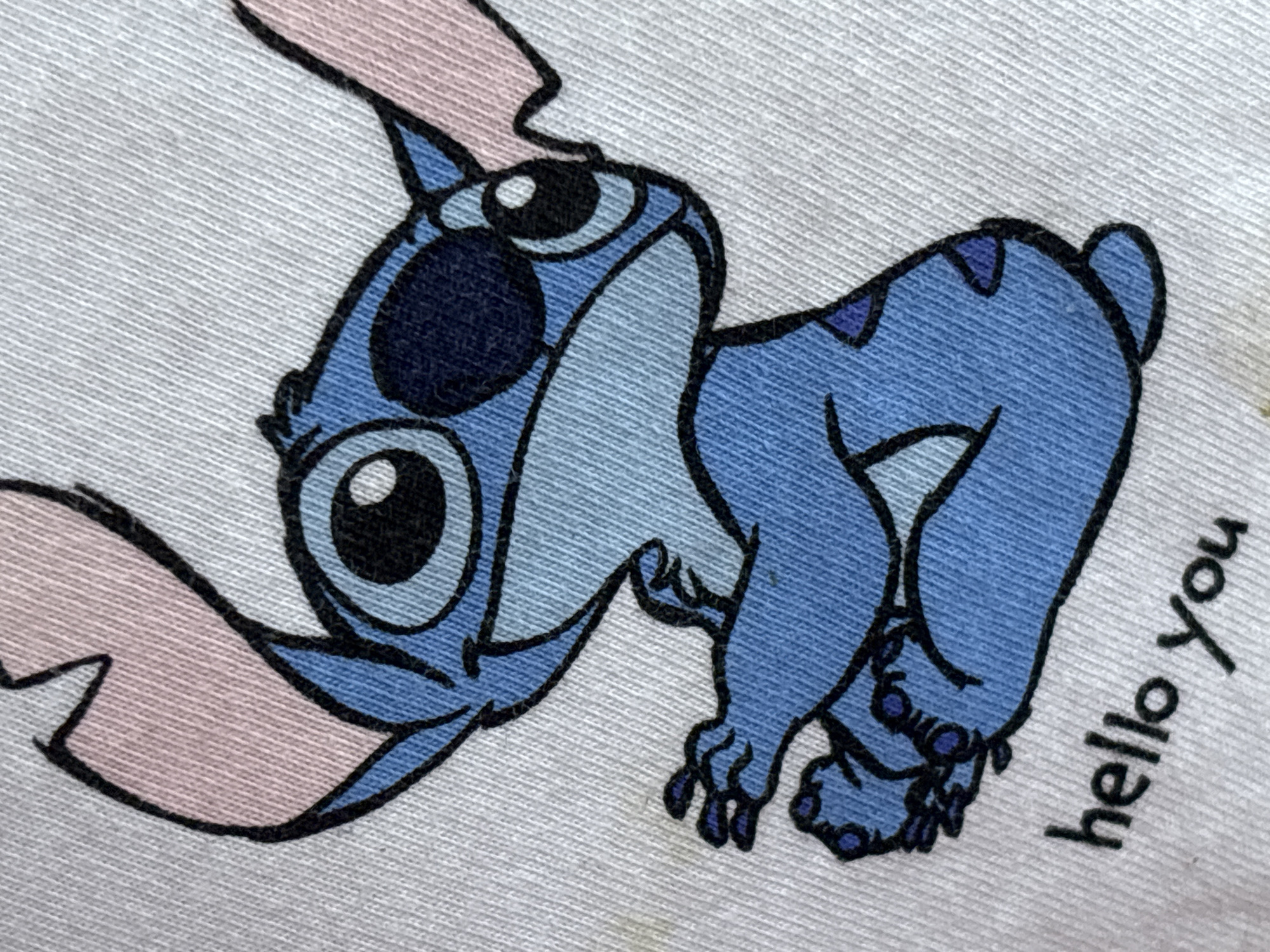 The image displays a close-up, slightly angled view of a white t-shirt featuring a cartoon illustration of the character Stitch from Disney's Lilo & Stitch. Stitch is depicted in blue with pink ears and a dark blue eyepatch, sitting with his arms crossed over his chest. Below the character, the words "hello you" are printed in black, stylized font. The t-shirt fabric shows a visible texture of woven threads. There is no discernible information in the image that places it in the city of Enevo, Bulgaria.