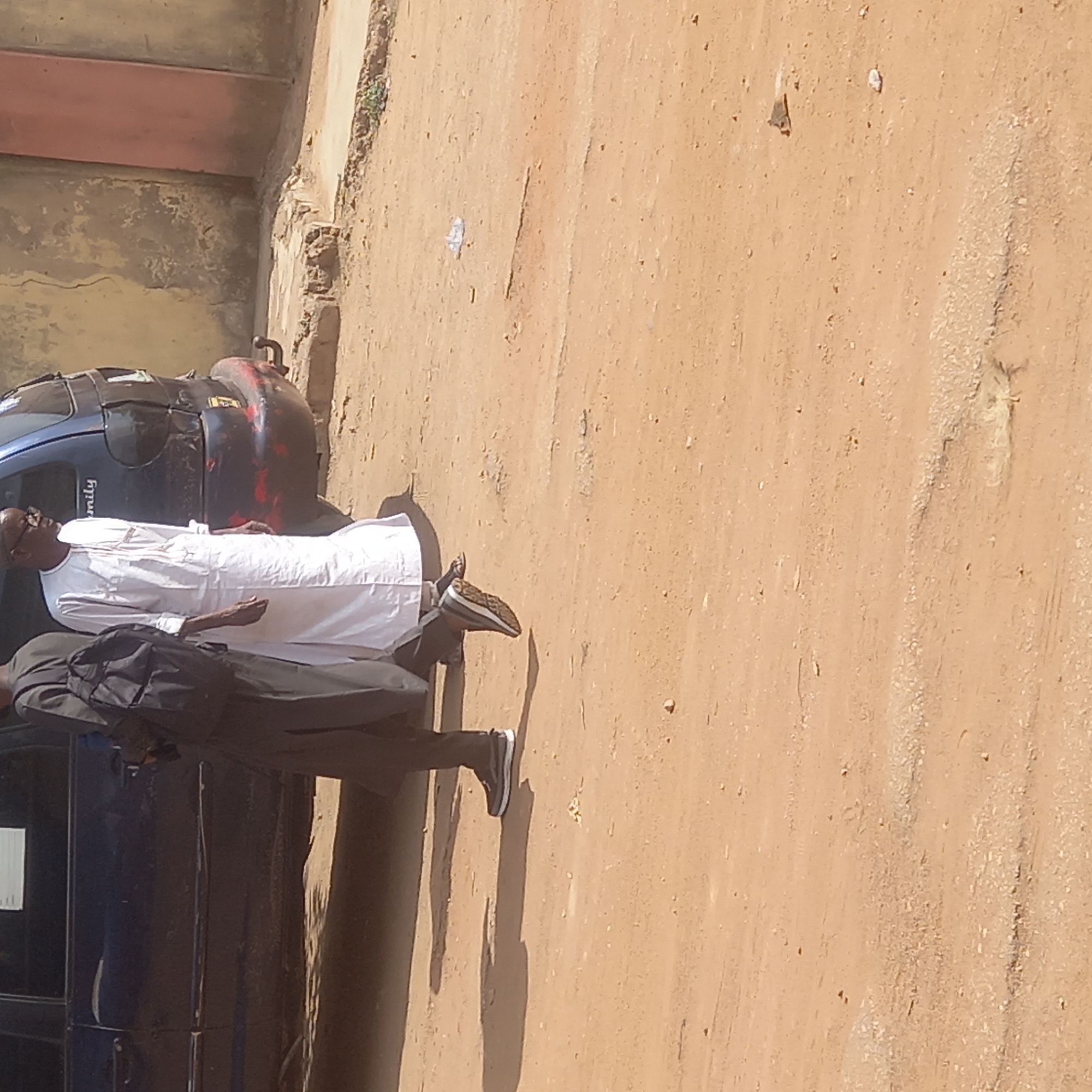 The image shows two men standing next to a blue car in Dadi Riba, Nigeria. One man is wearing a white robe and the other is wearing a black suit. The background is a dirt road and a building.
