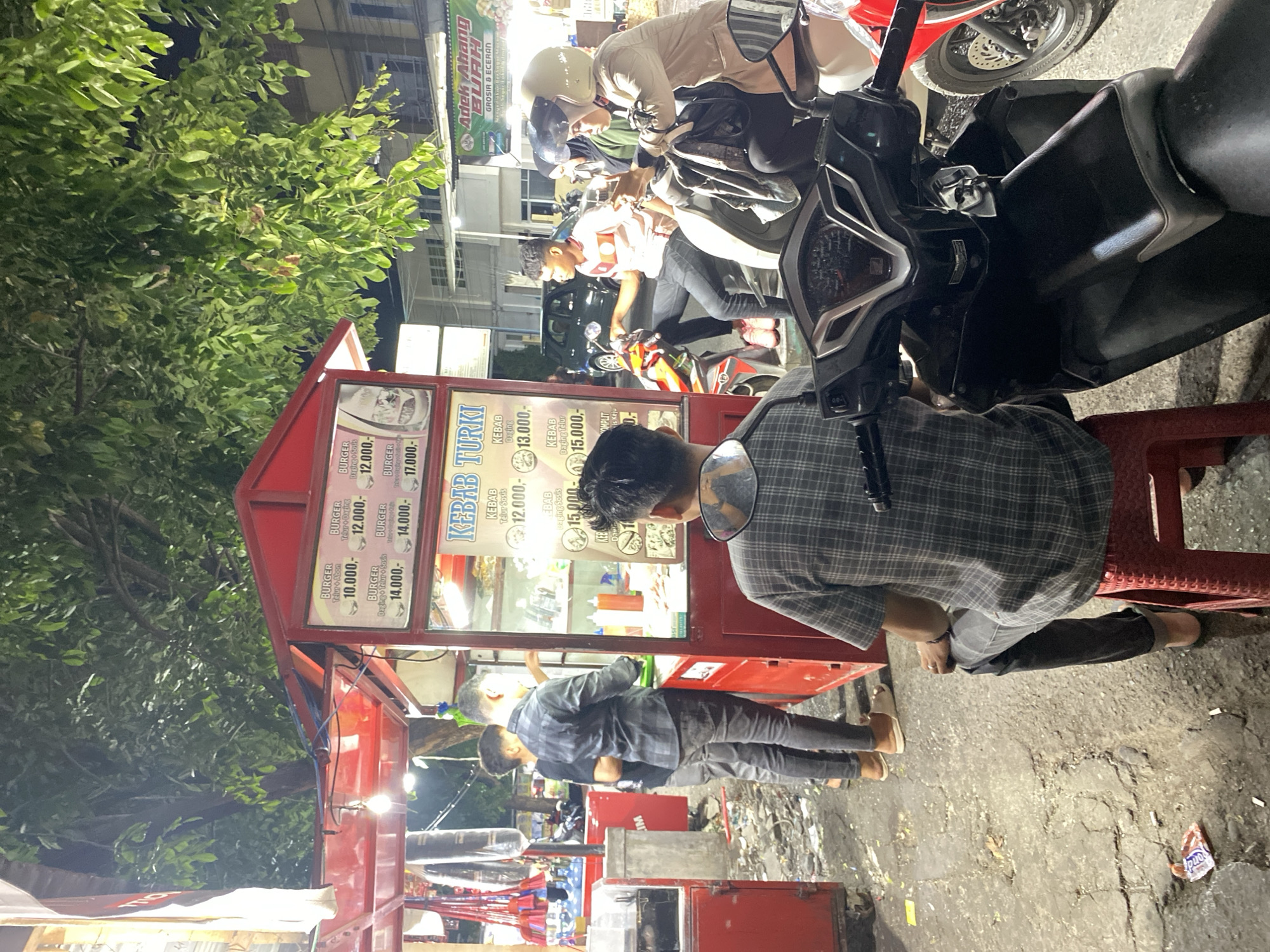 In Banda Aceh, Indonesia, at night, a street food stall selling burgers and kebabs is in operation. The stall features a red frame with illuminated signs displaying prices. Several individuals are present. Two men stand near the stall, one in a grey plaid shirt and black jeans, seemingly ordering. Another man, wearing a dark plaid shirt and grey jeans, is seated on a red plastic stool in the foreground, facing away from the camera. In the background, two people on a motorcycle, one wearing a helmet, are interacting. The ground is wet and littered, suggesting recent rain. Trees overhang the scene, and a building with a sign that reads "AdeK Abang BURAH GROSIR & ECERAN" is visible in the distance. The overall environment is that of a busy, informal street food setting.