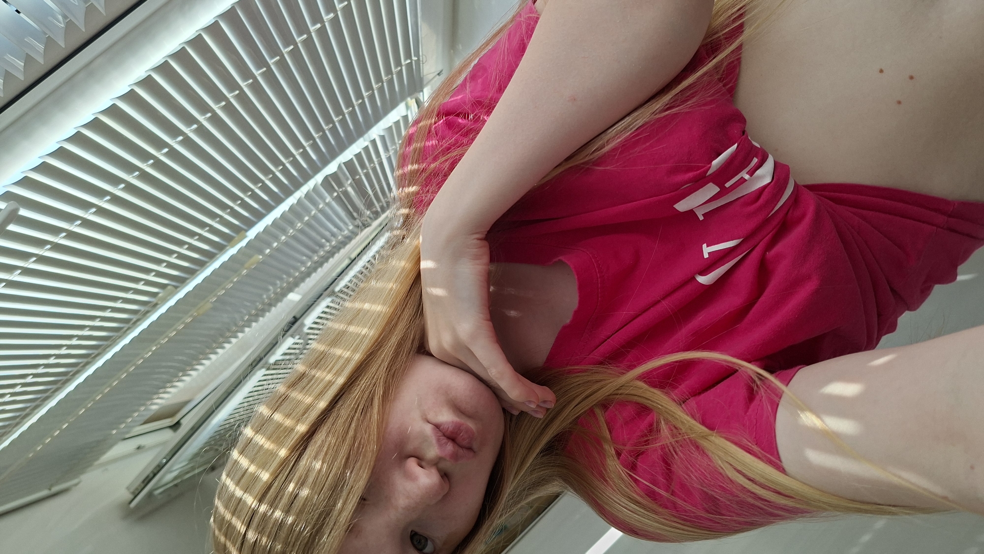 A woman with long, blonde hair and fair skin is posing indoors, next to a window with white horizontal blinds. She's wearing a bright pink t-shirt with white lettering. Her face is close to the camera, and one hand is near her face. The location is not discernible as Chaikovsky, Russia, from the image.