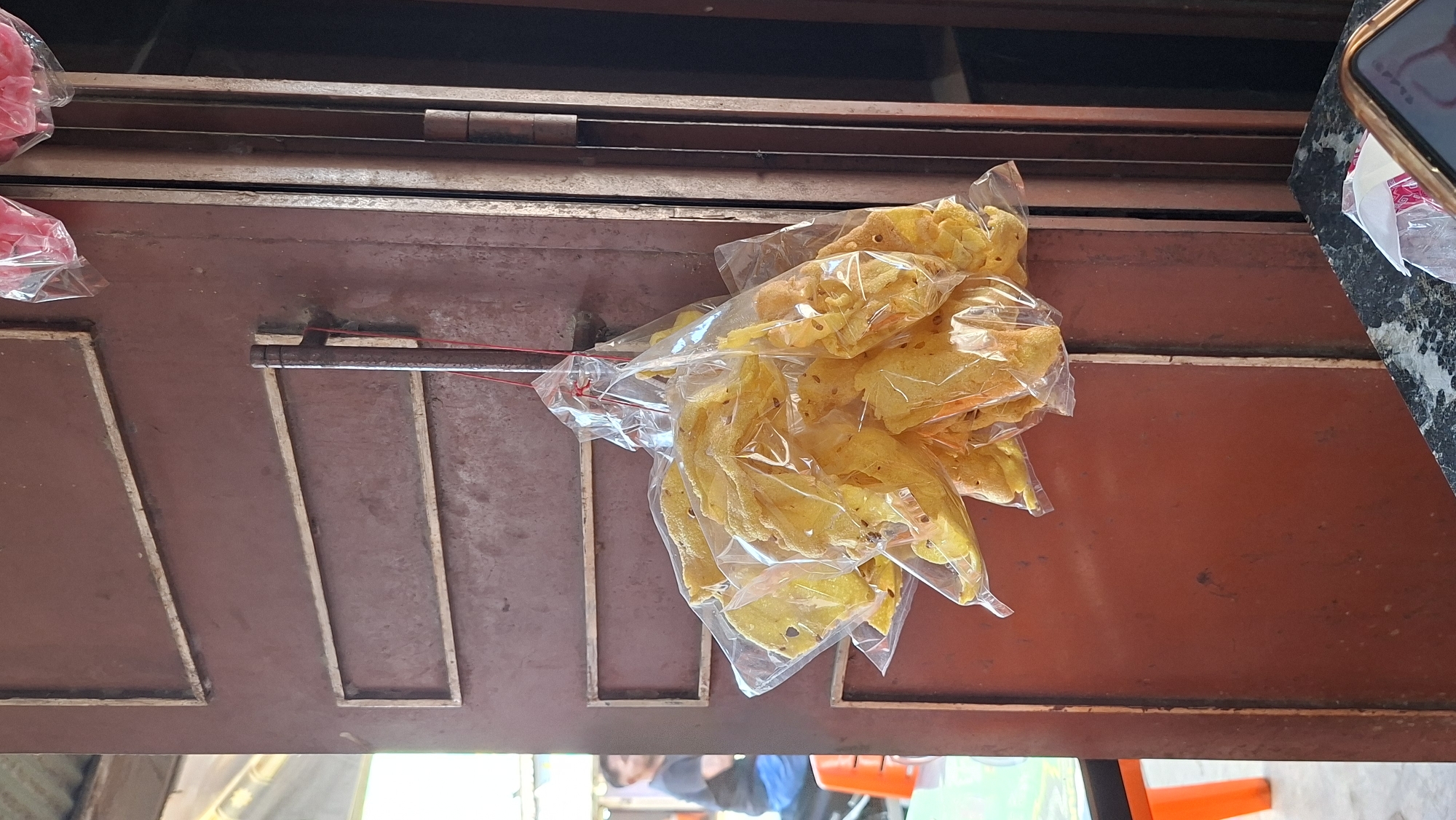 A bunch of transparent plastic bags filled with yellowish crackers are hanging from a metal hook attached to a reddish-brown metal door. The crackers appear to be thin and irregularly shaped, with some visible seeds within them. The door has two horizontal rectangular wooden frames on its surface. In the background, a person is visible sitting on an orange chair with a blurred background suggesting an outdoor or open-air setting, possibly a street or shopfront. The image is taken from a slightly low angle, looking up towards the hanging crackers. The overall scene suggests a street vendor or small shop in Keudebayu, Indonesia.