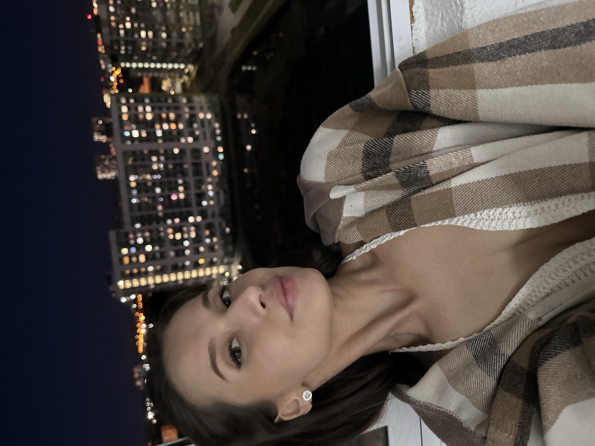 A woman is in the lower right of the image, in a vertical format. She has fair skin, dark eyebrows, and light pink lips. She is wearing a white top and a brown and white plaid blanket or shawl is draped over her shoulders. She is wearing a single diamond earring in her left ear. Behind her and to the left is a nighttime city scene with lit buildings. Based on the appearance of the building and the city lights, the location is consistent with a view of Yekaterinburg, Russia at night, but further confirmation would be needed for certainty. There are no visible actions or interactions occurring in the image.