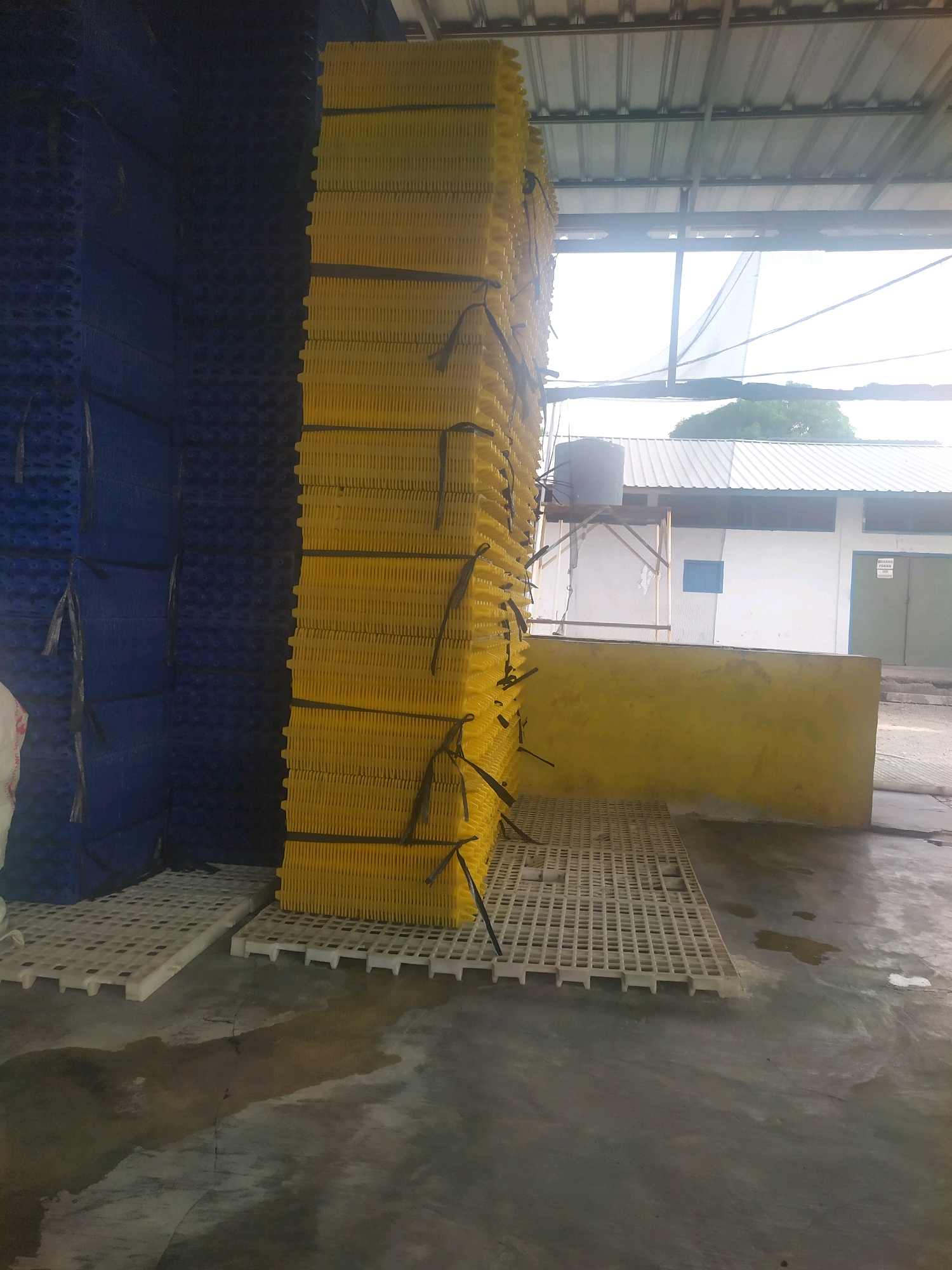 The image shows a stack of yellow crates and a stack of blue crates, likely in a storage or industrial area in Winong, Indonesia. The crates are made of plastic and are stacked high, secured with black straps. The ground is concrete and appears wet in some areas. In the background, there is a building with a corrugated metal roof and a yellow wall. A water tank is visible on a metal structure attached to the building. The overall scene suggests an outdoor or semi-outdoor storage facility. There are no people visible in the image.
