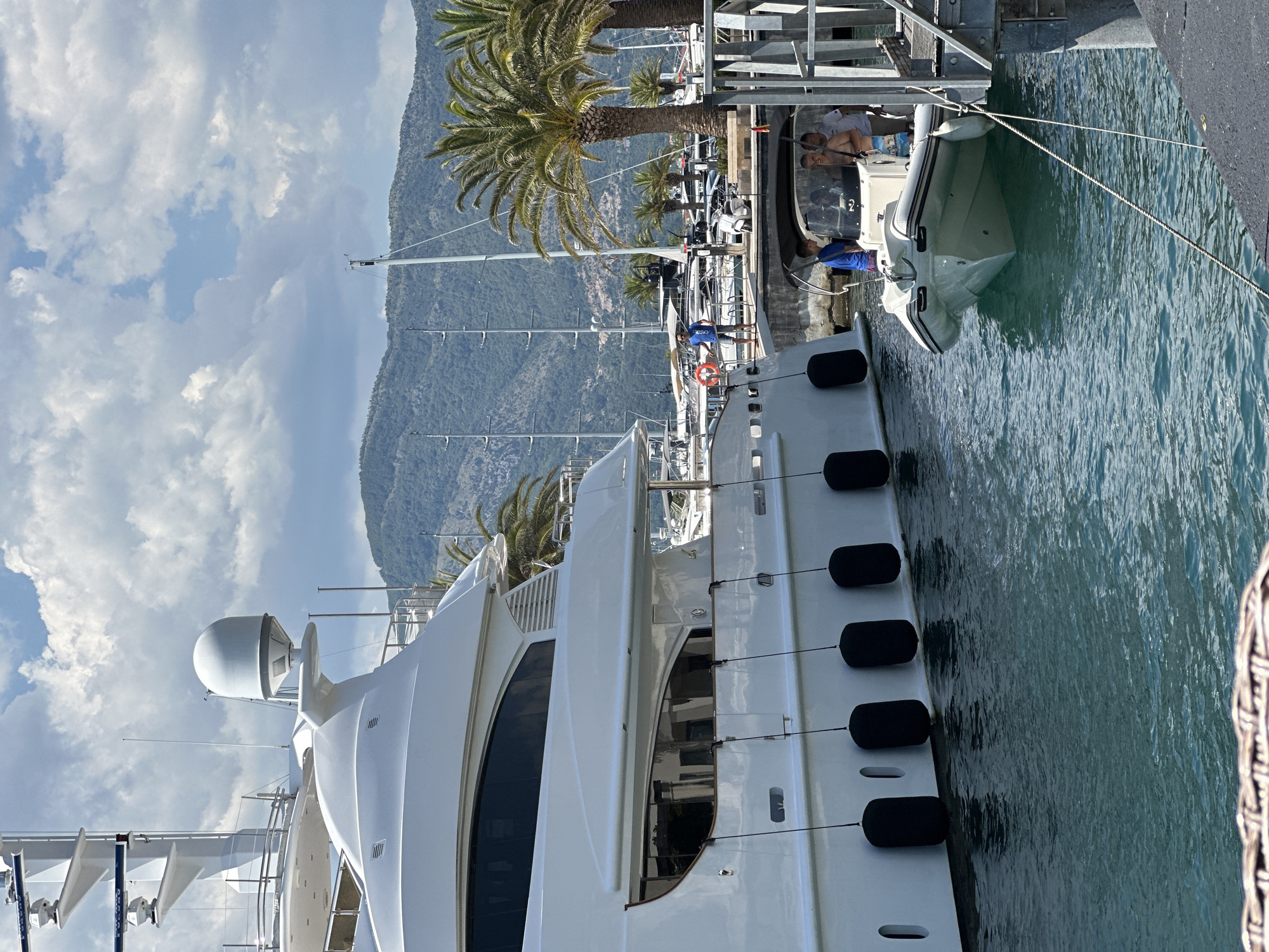 In Donja Lastva, Montenegro, a large white yacht is moored at a marina, its side featuring black fenders. Adjacent to it, a smaller white inflatable boat with a black canopy is docked, with three individuals visible inside and on the boat. The water is a clear turquoise. In the background, palm trees line the shore, and a mountainous landscape with a cloudy sky forms the backdrop. Several other yachts and sailboats are docked in the marina.