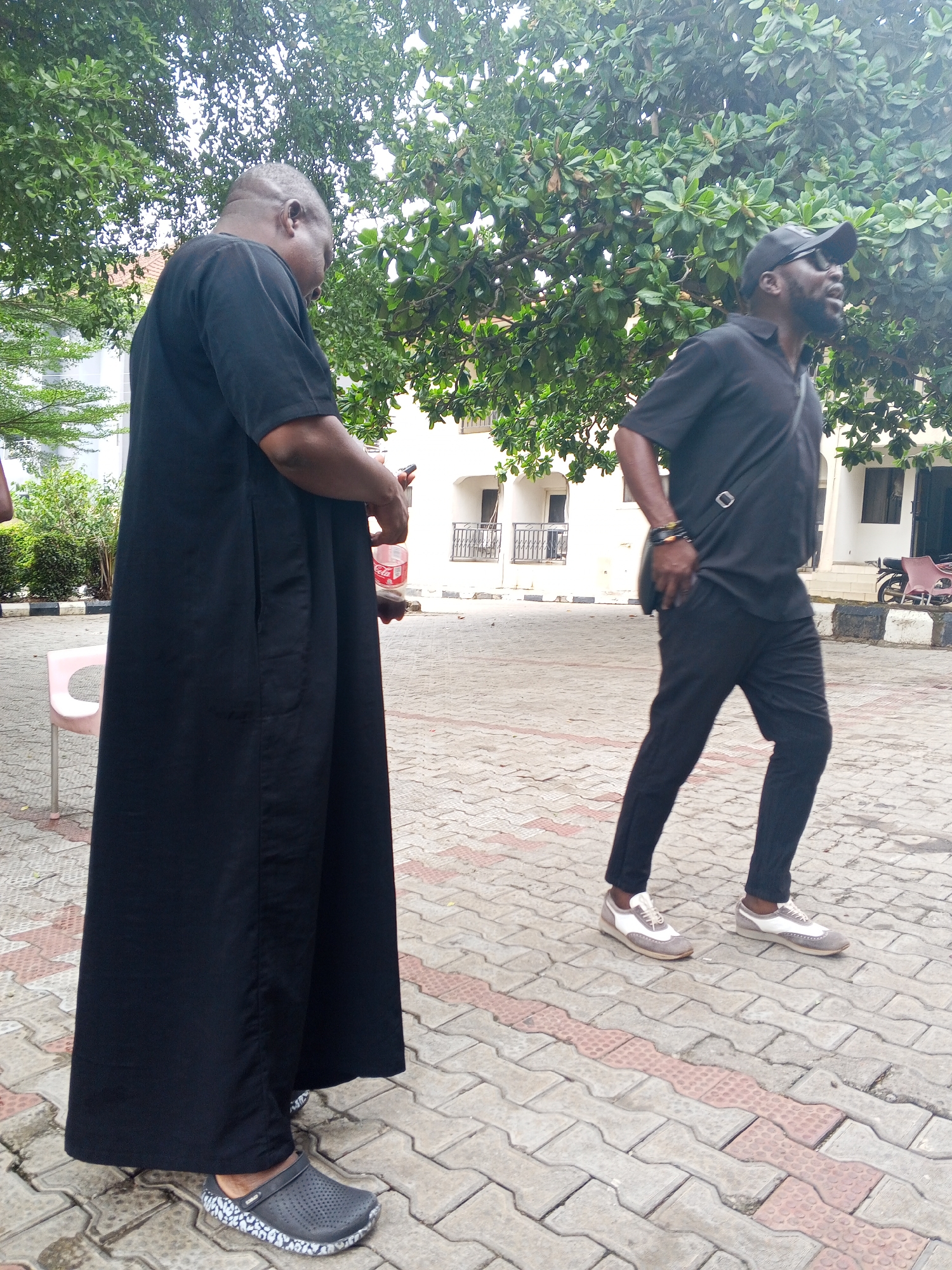 Two men are in an outdoor area paved with interlocking blocks, in Lokoja, Nigeria. The man to the left is wearing a long black robe and gray/blue Crocs. He appears to be holding a plastic bottle. The man to the right is wearing a black shirt, black pants, black baseball cap, a shoulder bag, and gray and white sneakers. He has a beard and sunglasses on top of his head and is gesturing with his mouth open. There are trees and buildings in the background. A pink chair is slightly visible behind the man on the left.