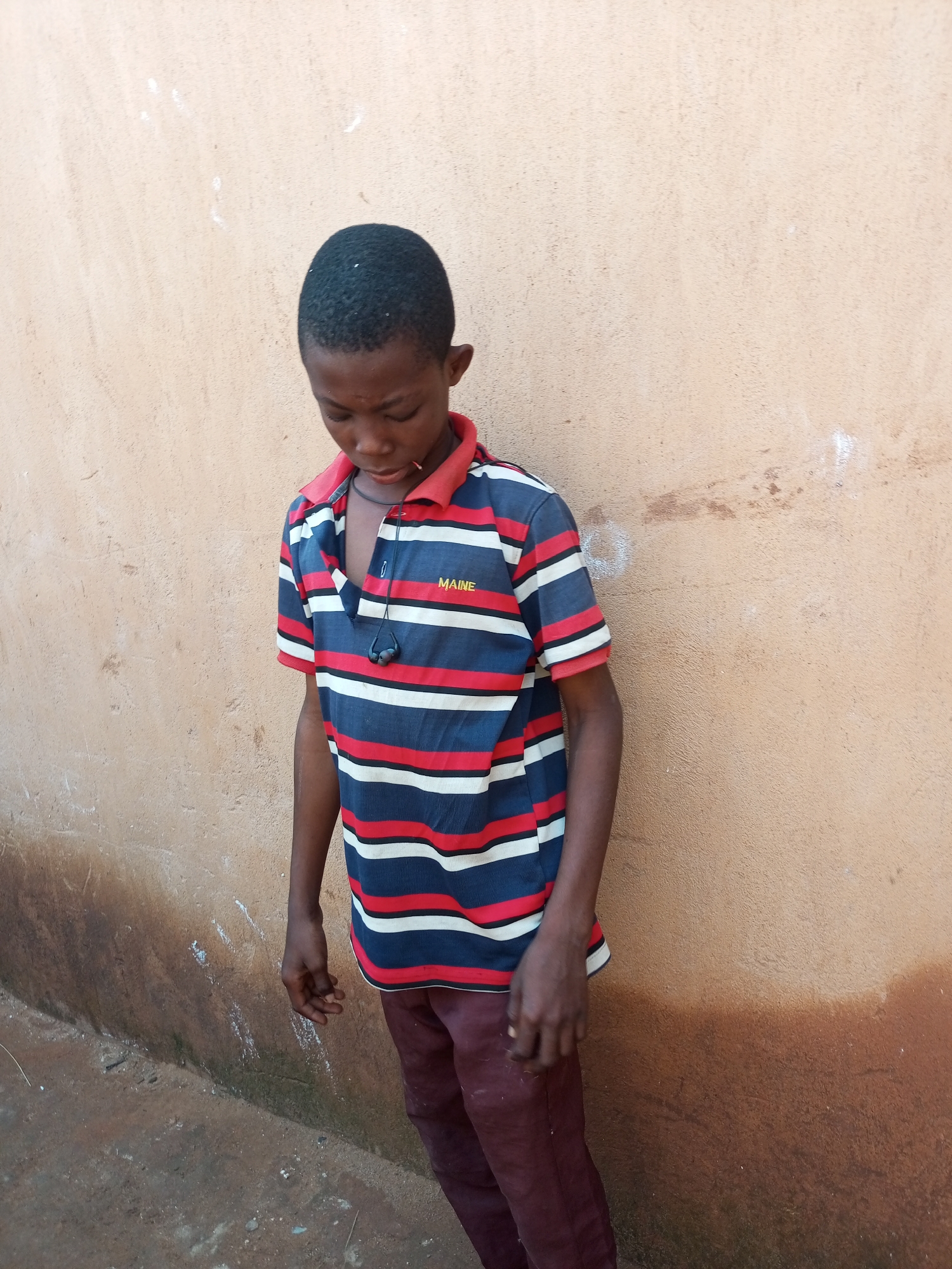 In Tempure, Nigeria, a young boy with dark skin and short hair stands in front of a textured, light brown wall. He is wearing a horizontally striped polo shirt with alternating bands of dark blue, red, and white. The shirt has a red collar and a small embroidered yellow text that appears to read "MAINE" on the right side of his chest. A black cord with two small earbud-like attachments hangs around his neck, resting on his shirt. He is also wearing dark maroon or burgundy pants. His head is bowed, and his gaze is directed downwards. His left hand is visible, with fingers slightly curled, near his waist. The wall behind him shows some subtle discoloration and marks. The ground in front of the wall is rough and appears to be a mix of dirt and possibly some debris.