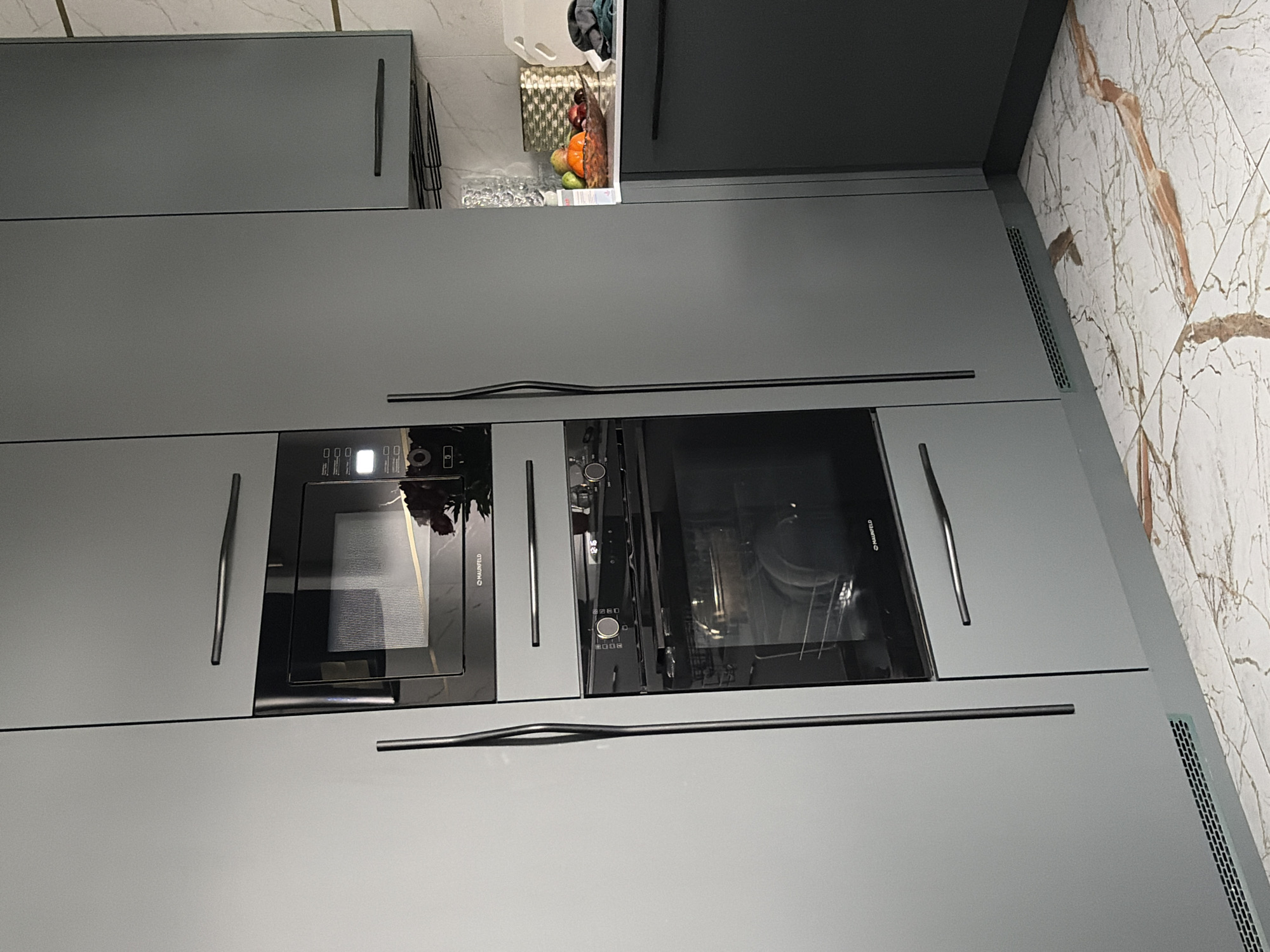 The image shows a modern kitchen with sleek, grey cabinetry. Integrated appliances, including a microwave and an oven, are visible within the cabinetry. The microwave is positioned above the oven, and both feature dark glass fronts. The oven door is slightly ajar, revealing racks inside. Long, vertical handles adorn the cabinet doors. The floor is tiled with a white and brown marble pattern. To the right of the main cabinetry, a section of a dark grey cabinet is visible, with a bowl of fruit on the countertop above it. The overall scene depicts a contemporary kitchen interior, likely a residential setting. The location is a kitchen in the city of Moscow, Russia.