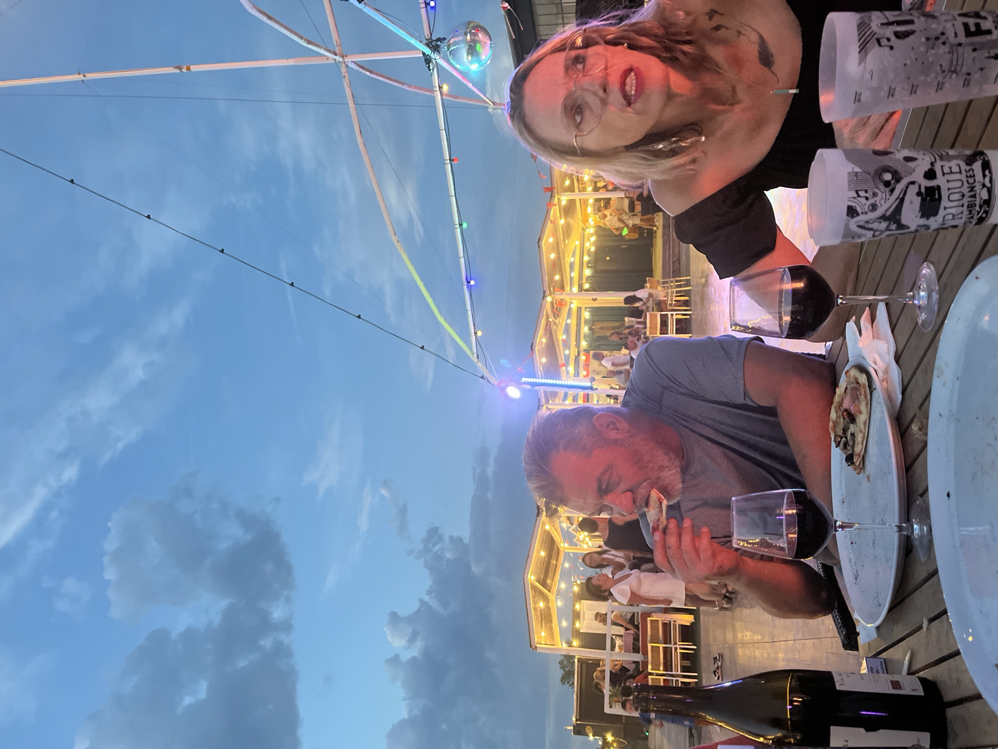 The image captures an outdoor dining scene under a twilight sky with dramatic clouds. In the foreground, a man with graying hair and a beard is seated at a wooden table, eating a slice of pizza. To his right, a woman with blonde hair and glasses is also seated at the table, looking away from the camera. In front of them are glasses of red wine, a bottle of wine, and plates with remaining pizza. The table is set on a paved area, and in the background, there are structures with illuminated string lights, creating a festive atmosphere. People can be seen dining and socializing at tables under these covered structures. The location is identified as Montélimar, France.