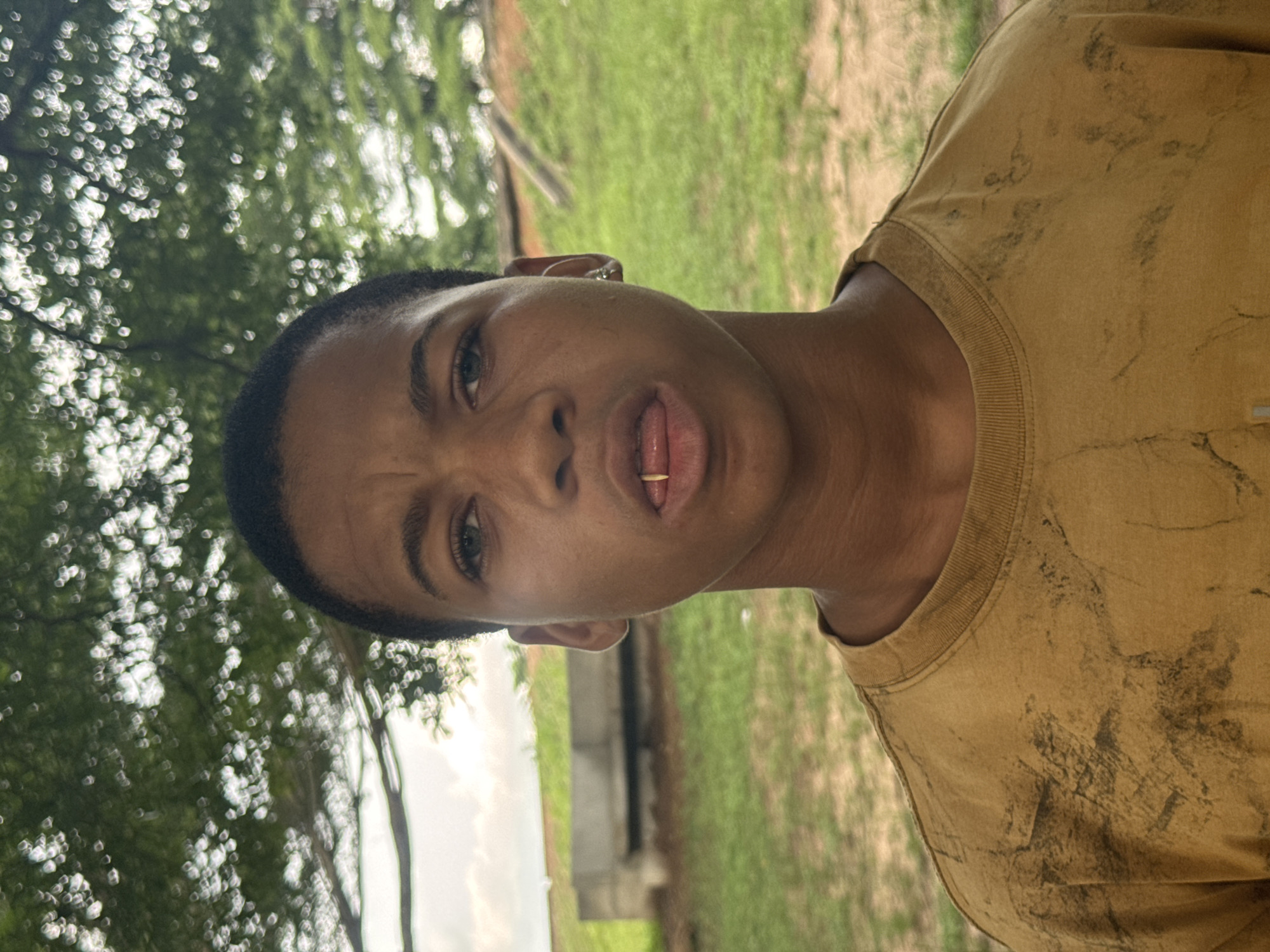 A medium close-up photograph shows a young, dark-skinned person wearing a mustard-colored t-shirt in Ihiagwa, Nigeria. The person’s face is expressive, with the mouth slightly open and a small object visible between the teeth. The subject appears to be outdoors in a grassy area with trees visible in the background. Other objects are obscured by the focus on the person.