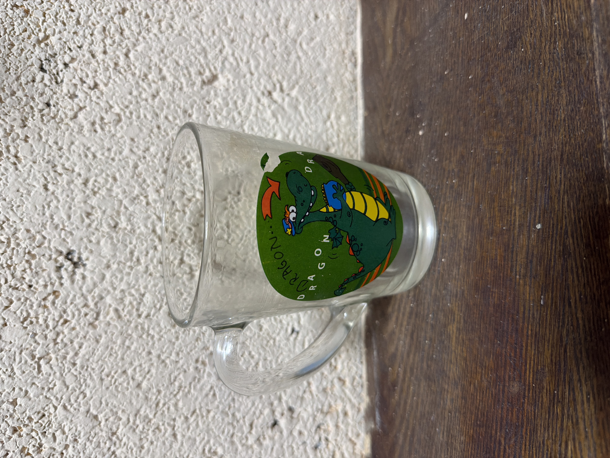 A clear glass mug is depicted lying horizontally on its side, bridging two distinct surfaces. The left portion of the mug rests on a light-colored, highly textured surface, consistent with a stucco wall or ceiling. The right side of the mug is positioned on a dark brown wooden surface, exhibiting prominent vertical grain, likely a floor or table.
The mug features a vibrant cartoon design on its exterior. Central to the design is a green dragon with a yellow underbelly and small orange accents. The dragon has large, wide eyes and appears to be interacting with a blue object. Surrounding the dragon, partially in an arc, the word "DRAGON" is visible in green lettering with white outlines. A small section of the green design appears to be chipped or faded. The mug itself is transparent glass, with a simple, C-shaped glass handle attached.
The background surfaces show minor specks and dust. The lighting suggests an indoor environment, casting subtle shadows and highlights on the mug and its surroundings. No people or specific activities are taking place; the image is a still life of the mug. The location and time of day are not discernible from the visual information.