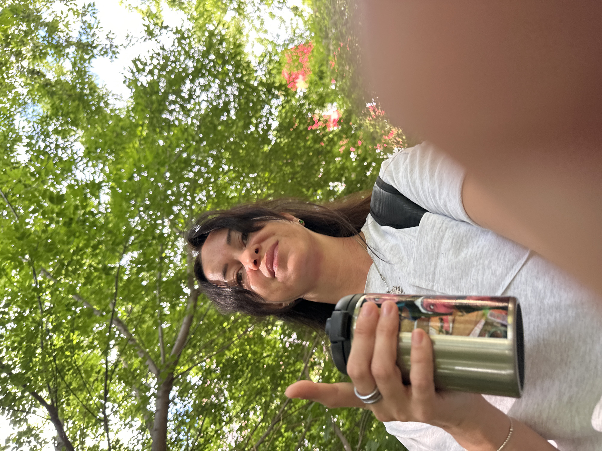 A low-angle, close-up shot captures a woman holding a reusable metallic cup while standing outdoors, with lush green foliage overhead and dappled sunlight. She is wearing a light-colored t-shirt with a dark backpack strap visible on her left shoulder. The woman has dark hair, a silver necklace, and a ring on her right index finger. The cup she holds is decorated with a colorful, graphic design. The environment suggests a park or wooded area, with blurred green leaves dominating the background and hints of pink flowers in the distance. There is no discernible urban setting or identifiable landmarks that would suggest the city of Irkutsk, Russia.