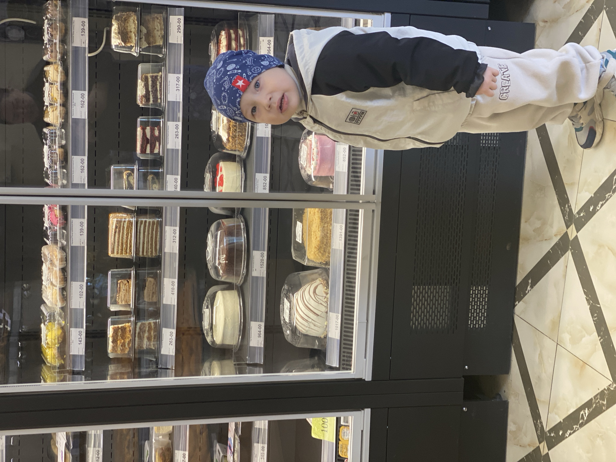 A young boy wearing a blue hat, black jacket, and white pants stands in front of a refrigerated display case filled with various cakes, likely in a bakery or grocery store in Sochi, Russia. The cakes are individually packaged and priced, with labels visible on the shelves. The boy is looking at the camera. The floor has a marbled pattern.