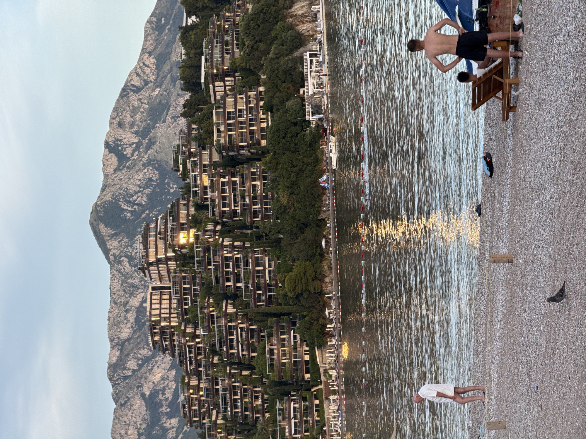 The image depicts a coastal scene at dusk, likely in Budva, Montenegro, given the distinctive terraced resort buildings cascading down the hillside. The resort, with its numerous balconies and windows, is situated against a backdrop of rugged mountains. In the foreground, a pebbled beach meets the calm sea, which reflects the golden light of the setting sun.
On the beach, several individuals are present. An older man in a white shirt and shorts walks along the shoreline. Further down the beach, two young men are visible. One stands with his hands on his hips, facing away from the camera, while another is seated on a lounge chair. A pigeon stands on the beach near a wooden post.
The water is marked by a floating line of buoys, separating the swimming area. A small white boat is anchored near the shore. The overall atmosphere is one of a peaceful evening at a seaside resort.