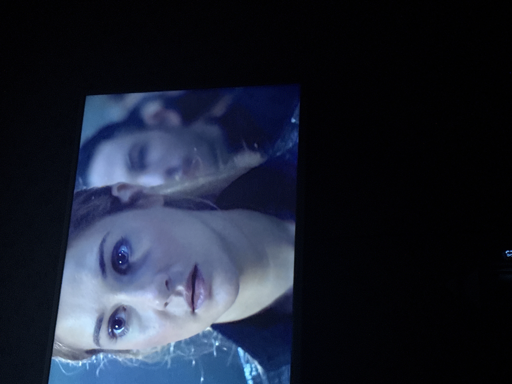 The image shows a close-up of two people's faces, oriented at a slight angle. In the foreground is a woman with fair skin, light hair, and blue eyes. Her expression appears to be one of surprise or concern. Behind her, partially obscured, is a man with dark hair, looking sideways, also with a somber expression. The background is dark and slightly blurred, suggesting the image is captured from a screen or monitor. The location is impossible to determine from the image.