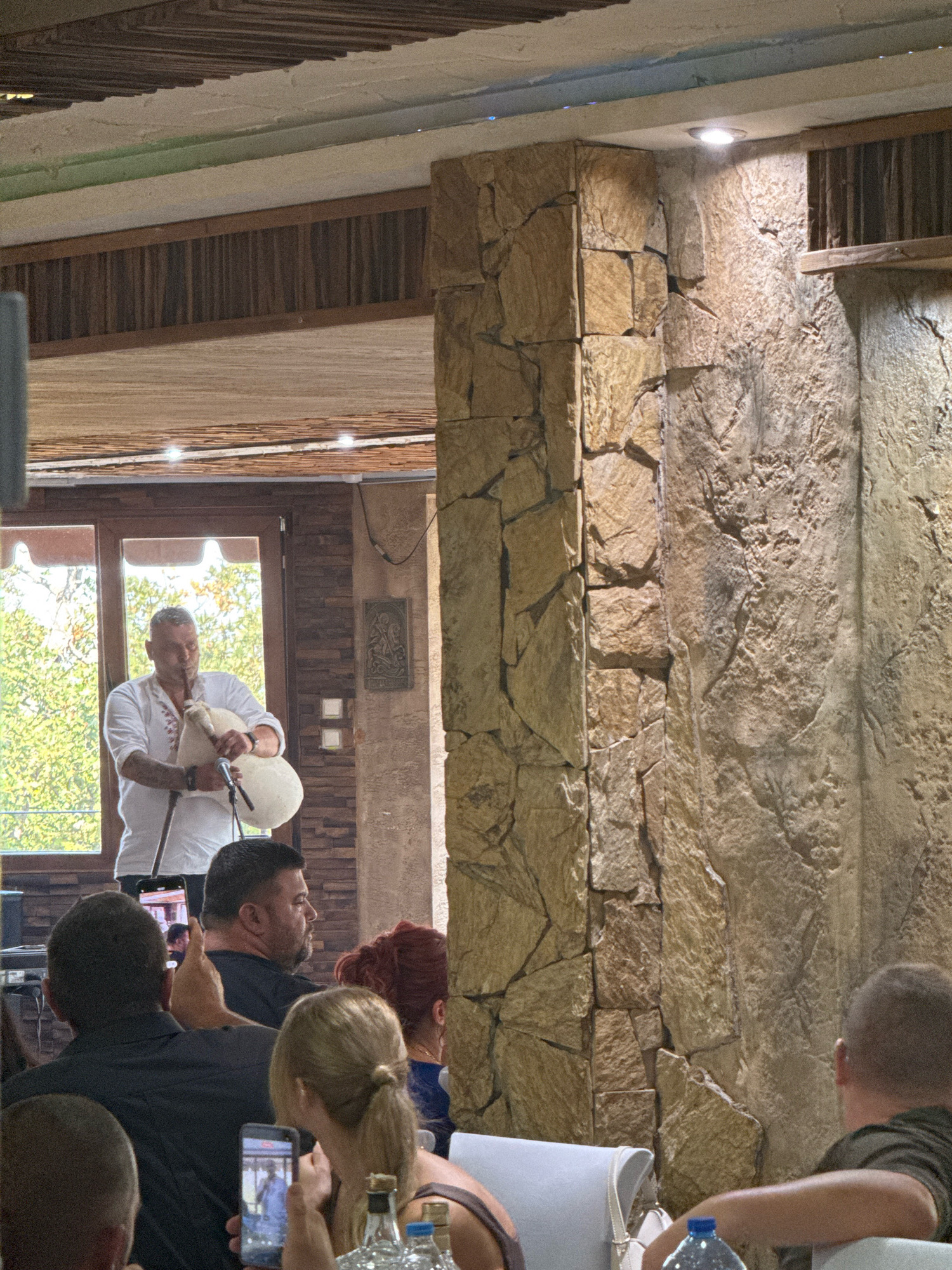 The image shows a man playing bagpipes in what appears to be an indoor venue. The man is wearing a white shirt with red embroidery. He is standing in front of a window with greenery visible outside. There are several people in the foreground, some of whom are holding up their mobile phones, possibly to record the performance. The venue has rustic stone pillars and wooden ceiling elements. The atmosphere suggests a cultural event or performance. The provided location information indicates the scene is in Sredets, Bulgaria.