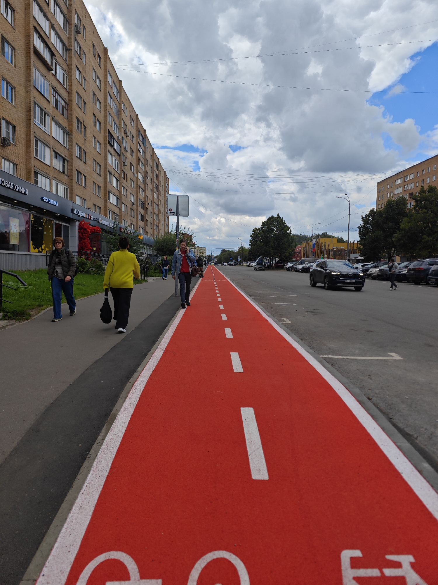 In Lobnya, Russia, on a cloudy day, a red-colored bike lane runs alongside a paved road. Several pedestrians are visible, including a person in a yellow sweater walking away from the camera on a sidewalk, and another person in a blue denim jacket walking towards the camera on the bike lane. Cars are parked along the road, and one car is driving in the lane next to the bike path. The scene is dominated by the bright red bike lane, with a tall brick apartment building on the left and trees and other buildings on the right. The sky is filled with large, dramatic clouds.