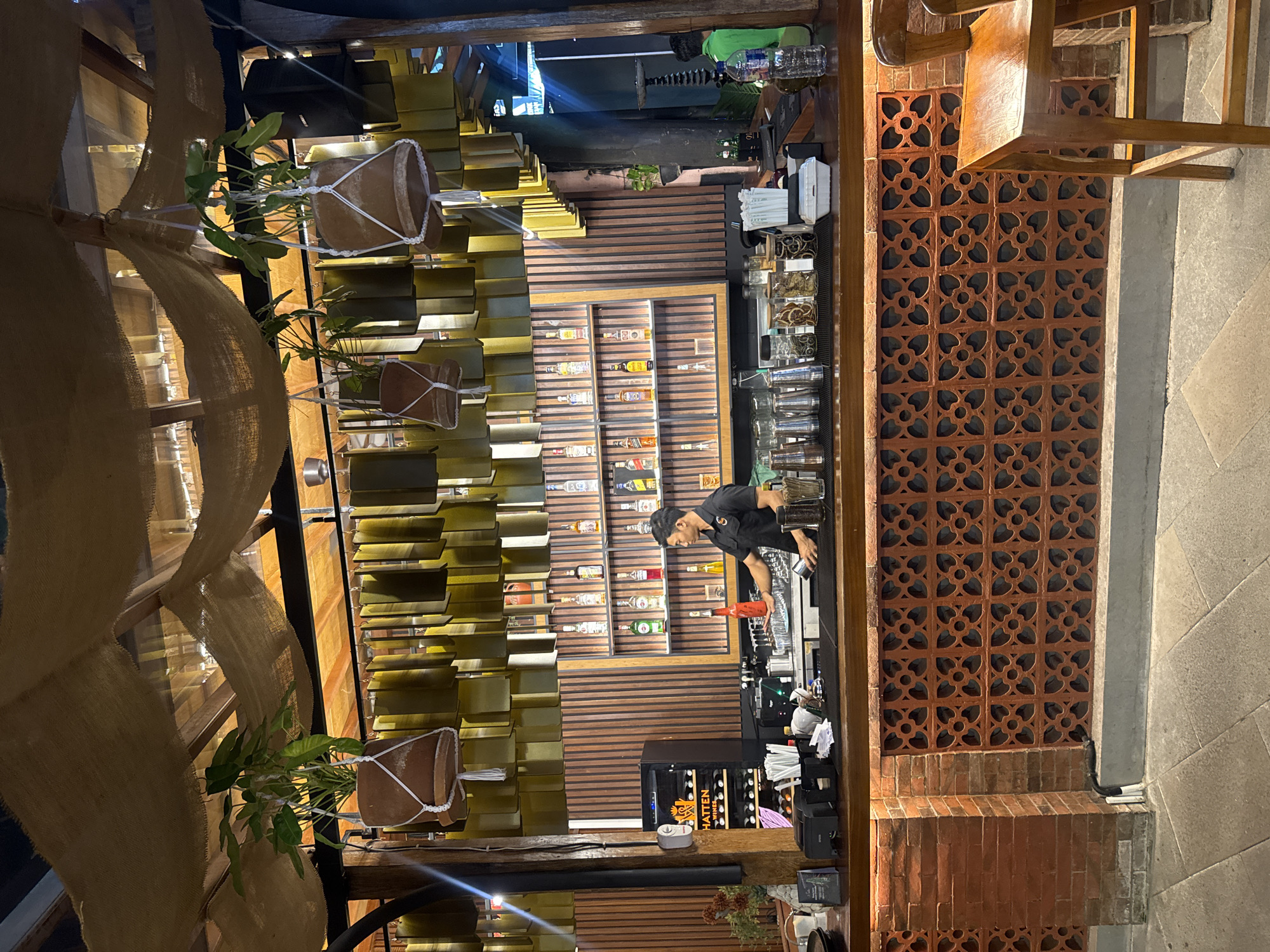 This image depicts the interior of a bar located in Kuta, Indonesia. The bar features a wooden counter with various bottles of alcohol displayed on shelves behind it. A bartender, dressed in a black polo shirt, is actively preparing drinks. Hanging plants in terracotta pots are suspended from the ceiling, adding to the ambiance. The walls are adorned with wooden paneling and decorative elements. A refrigerator unit, labeled "HATTEN WINES," is visible to the left of the bar. The floor is paved with stone tiles. The overall atmosphere suggests a relaxed and inviting establishment.
