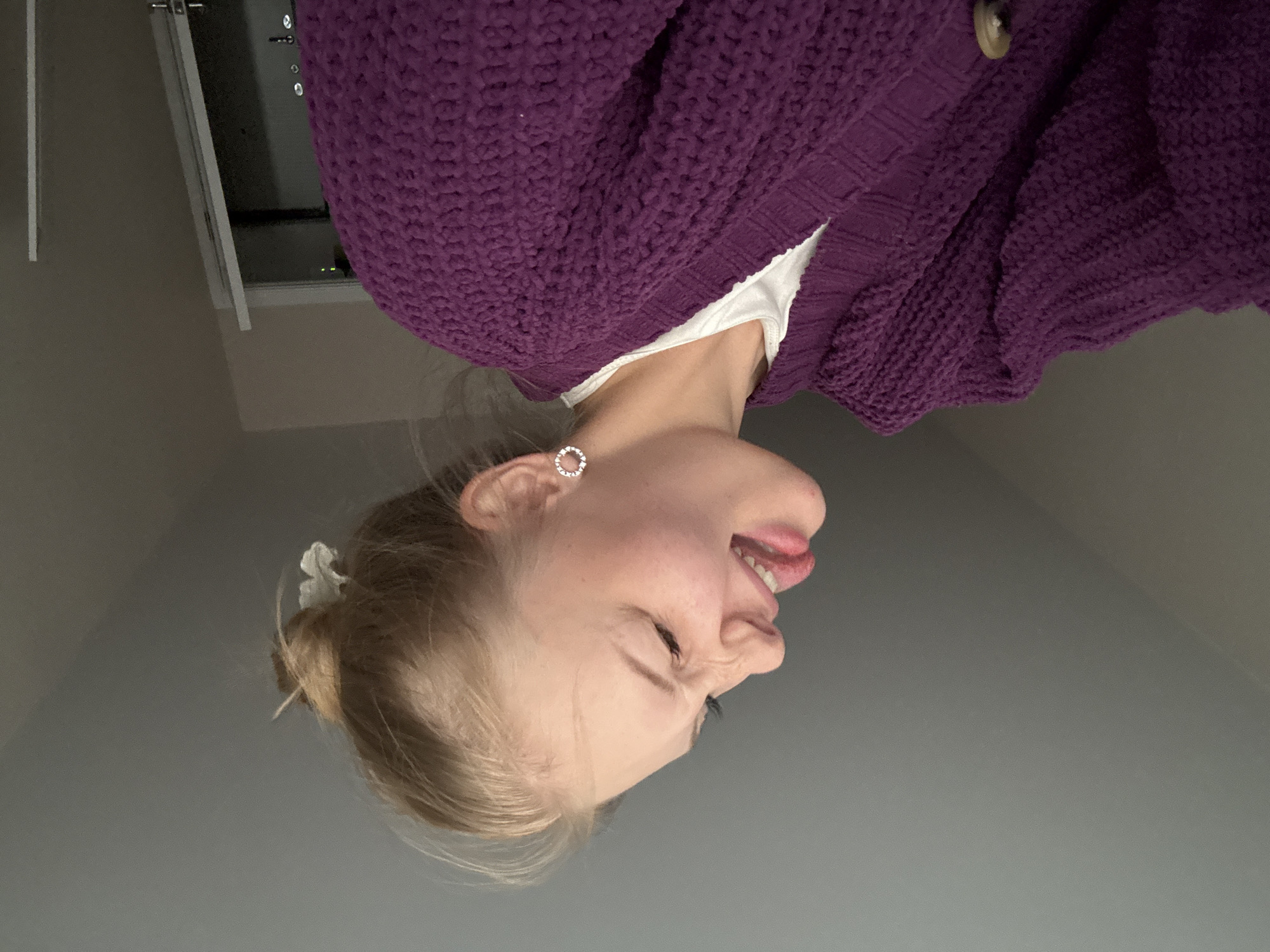 A young woman with blonde hair pulled back in a bun, wearing a purple chunky knit cardigan over a white t-shirt, sticks her tongue out and scrunches her nose while looking to her left, her eyes closed. She has a small, white flower clipped into her hair and wears a circular, rhinestone earring in her visible ear. The background appears to be an indoor setting with a grey ceiling and wall, and a doorway is visible on the right side of the frame. This image was taken on February 11, 2023, in Perm, Russia.