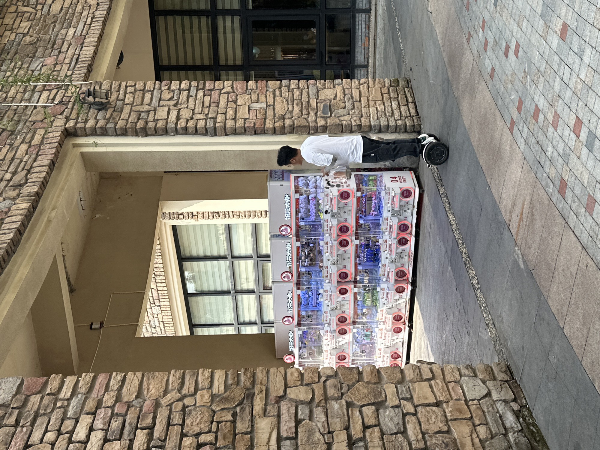 A person is using a gumball machine on a street in Chongqing High-tech Zone, China. The person is wearing a white shirt and dark pants, riding on a hoverboard or similar self-balancing scooter. The gumball machine stands between two sections of brick wall. The street is paved with concrete and tiles. A building with a window is visible in the background.