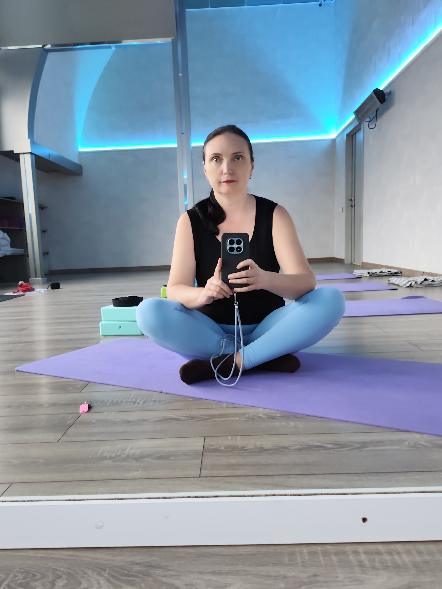 A woman with dark hair is sitting cross-legged on a purple yoga mat in a yoga studio. She is wearing a black tank top and light blue leggings. She is holding a black smartphone with multiple cameras in her hands, taking a selfie. The background of the studio features blue lighting along the ceiling and walls, wooden flooring, and other yoga mats laid out. In the background, there are shelves with folded white cloths and a door. The overall environment suggests a clean and modern fitness space.