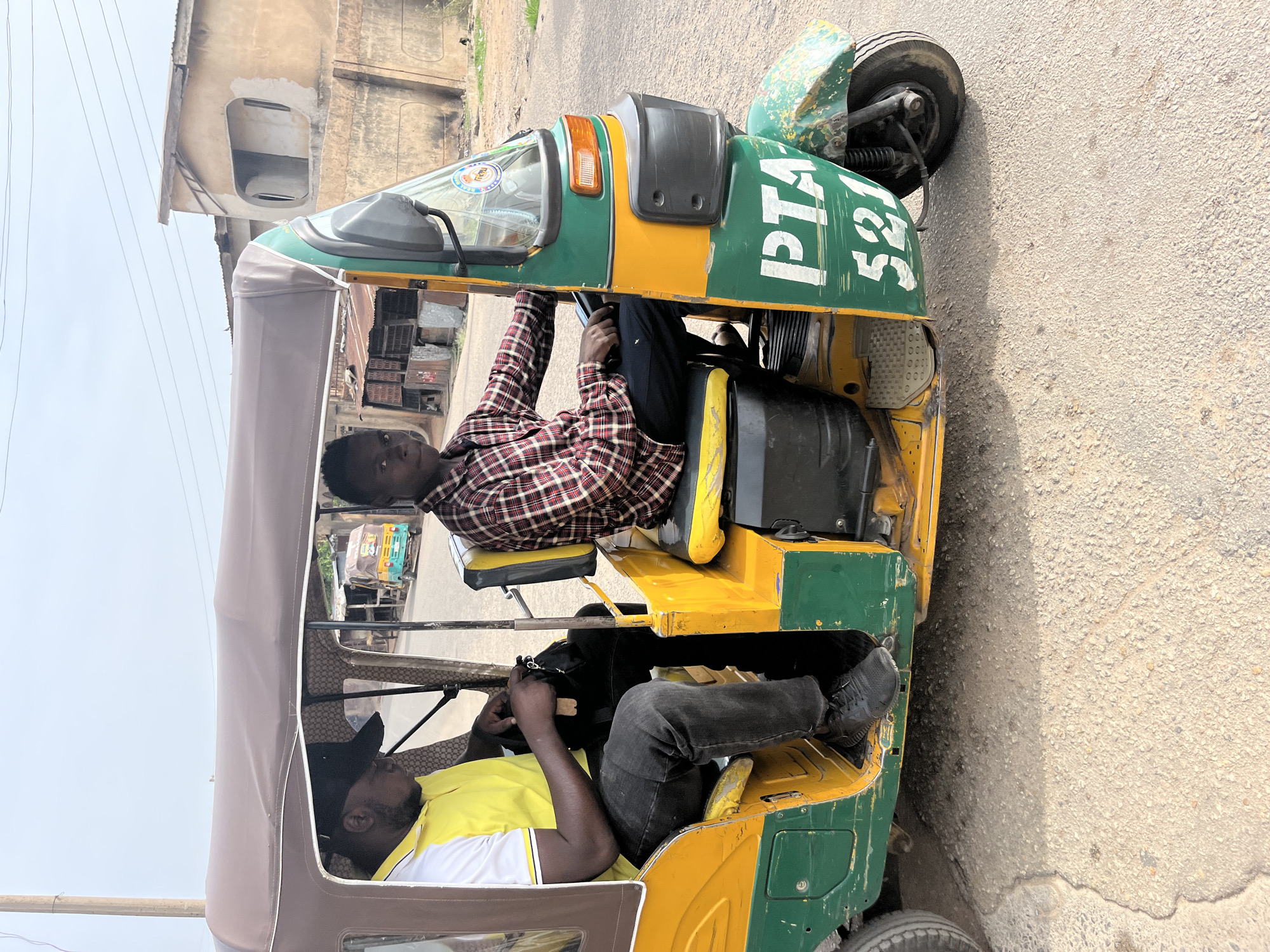 Two men are inside a green and yellow "keke napep" tricycle taxi on a paved road in Jos, Nigeria. The man in the driver's seat wears a yellow and white shirt and a black baseball cap, while the other man wears a red and black plaid shirt. The tricycle has the letters "PTA 521" painted on the front. Buildings are visible in the background.