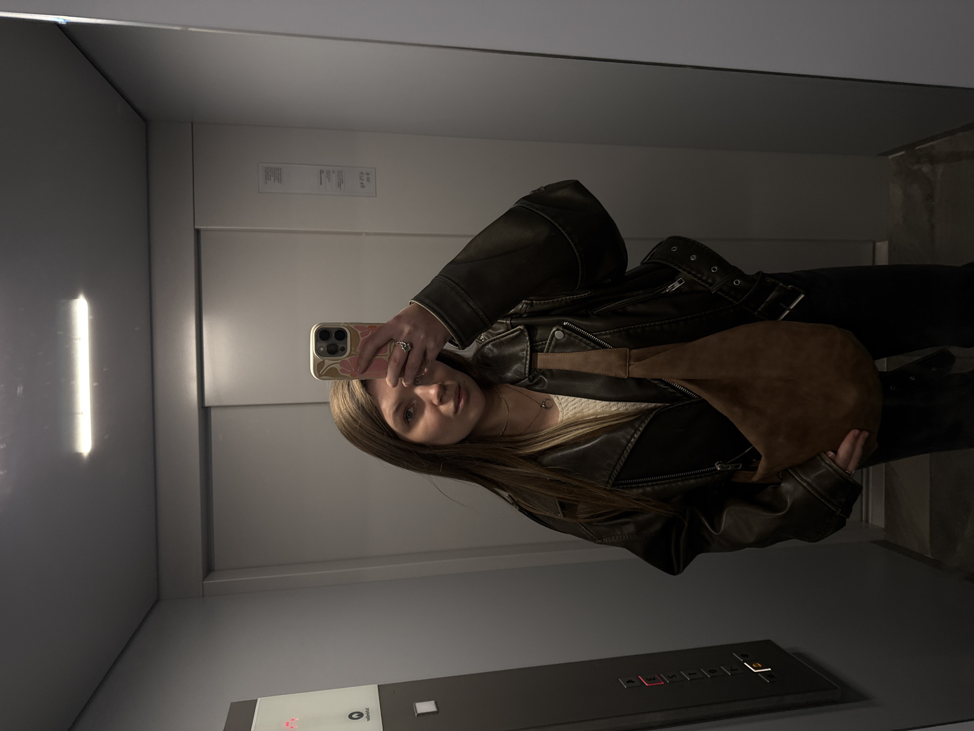A young woman with long brown hair is taking a selfie in a mirrored elevator. She is wearing a black leather jacket and holding a brown handbag. The elevator has grey walls and a control panel with illuminated buttons. The image was taken in an urban environment, likely in Vozdovac Urban Municipality, Serbia.