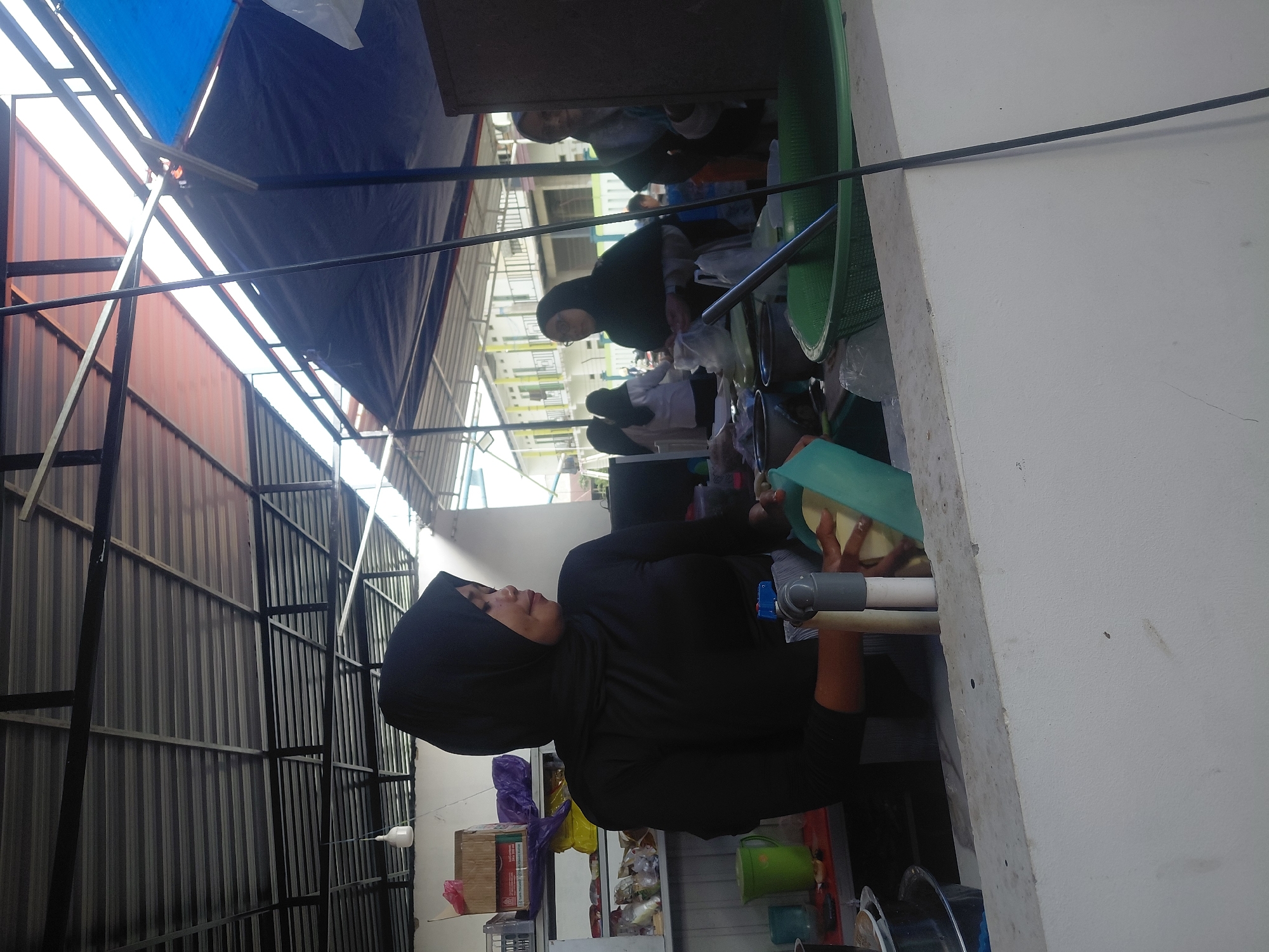 This image depicts an outdoor market stall, likely in Kendari, Indonesia, under a corrugated metal roof supported by metal beams. The stall appears to be a food vendor. A woman wearing a black hijab and a black shirt is the central figure, actively engaged in what looks like food preparation. She is holding a light green plastic colander or bowl in her left hand and appears to be washing or draining something. Her right hand is out of frame but seems to be assisting with the task.
To her left, there is a green pitcher and various items on a shelf, including a cardboard box and purple packaging. In front of her, a white concrete counter separates her from the viewer. Behind her, to the right, other individuals are visible, also appearing to be vendors or customers. One person in the background is wearing glasses and a hijab, and another person is partially obscured by a dark structure. The environment suggests a bustling, open-air marketplace with a mix of goods and daily commerce. Sunlight filters through gaps in the roofing, indicating it is daytime. The overall impression is one of a typical day at a local Indonesian market.