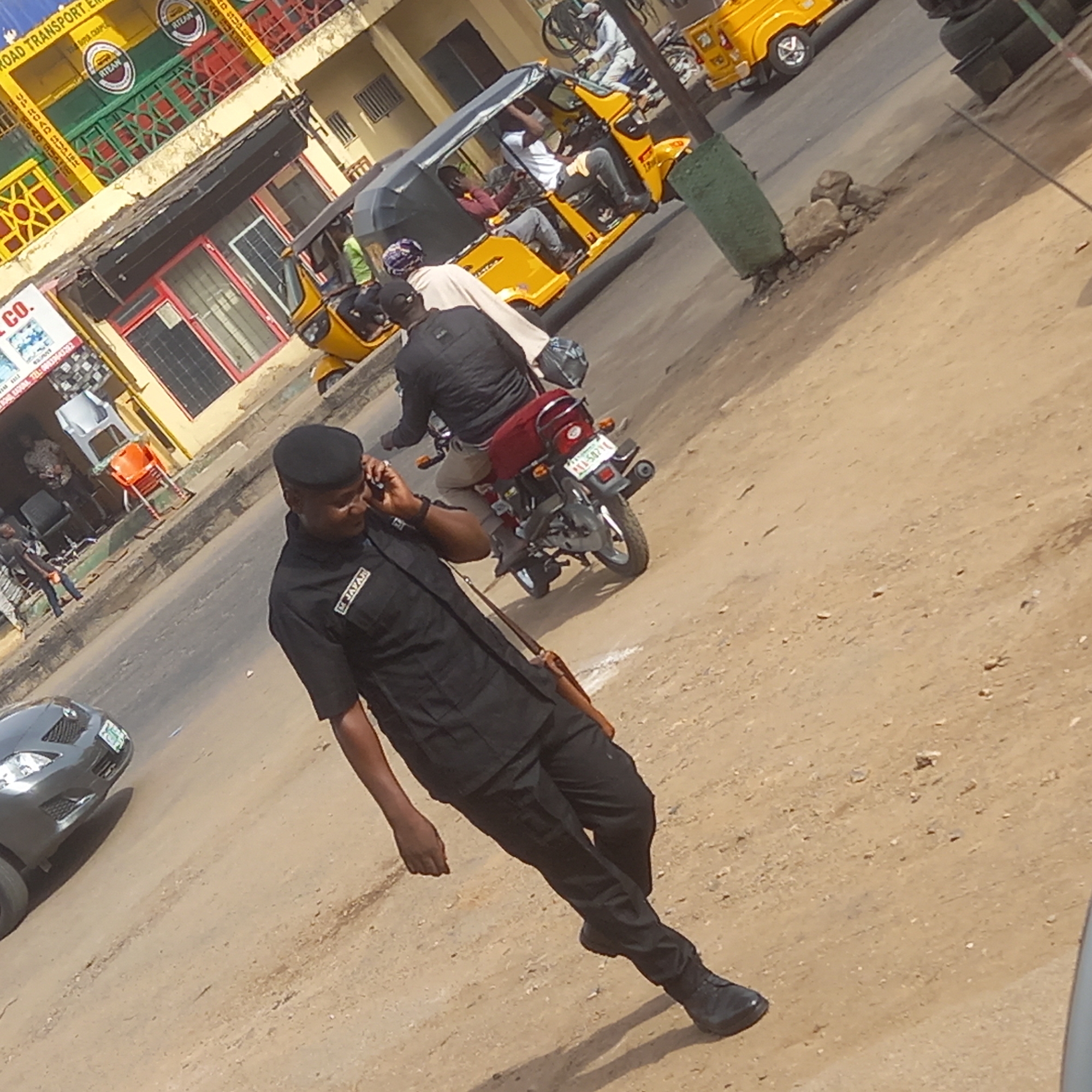 This image captures a bustling street scene in Dadi Riba, Nigeria, during broad daylight. The main subject is a man in the foreground, wearing a black uniform comprising a short-sleeved shirt, trousers, and a black cap. He is walking on a dusty, light brown road, holding a mobile phone to his right ear, seemingly in conversation. A name tag or patch on his left chest reads "M. T. JAZAH".
Behind the man, the street is active with vehicles. A person on a motorcycle, dressed in a black jacket, is driving away from the viewer. The motorcycle has a visible license plate: "YM 582YH". Further along the road, several yellow three-wheeled vehicles, commonly known as Keke Napeps, are seen, carrying passengers. Another motorcycle is also partially visible in the background.
To the left, commercial buildings line the street. One building features a distinctive multi-colored (yellow, green, red) facade with a sign that partially reads "ROAD TRANSPORT EM..." and displays a circular logo with the text "RTZAN" multiple times. Another sign below it has "CO." visible. The environment suggests an urban or semi-urban setting with typical daily activities and transportation. The road surface appears unpaved or heavily worn in areas, contributing to the dusty appearance.