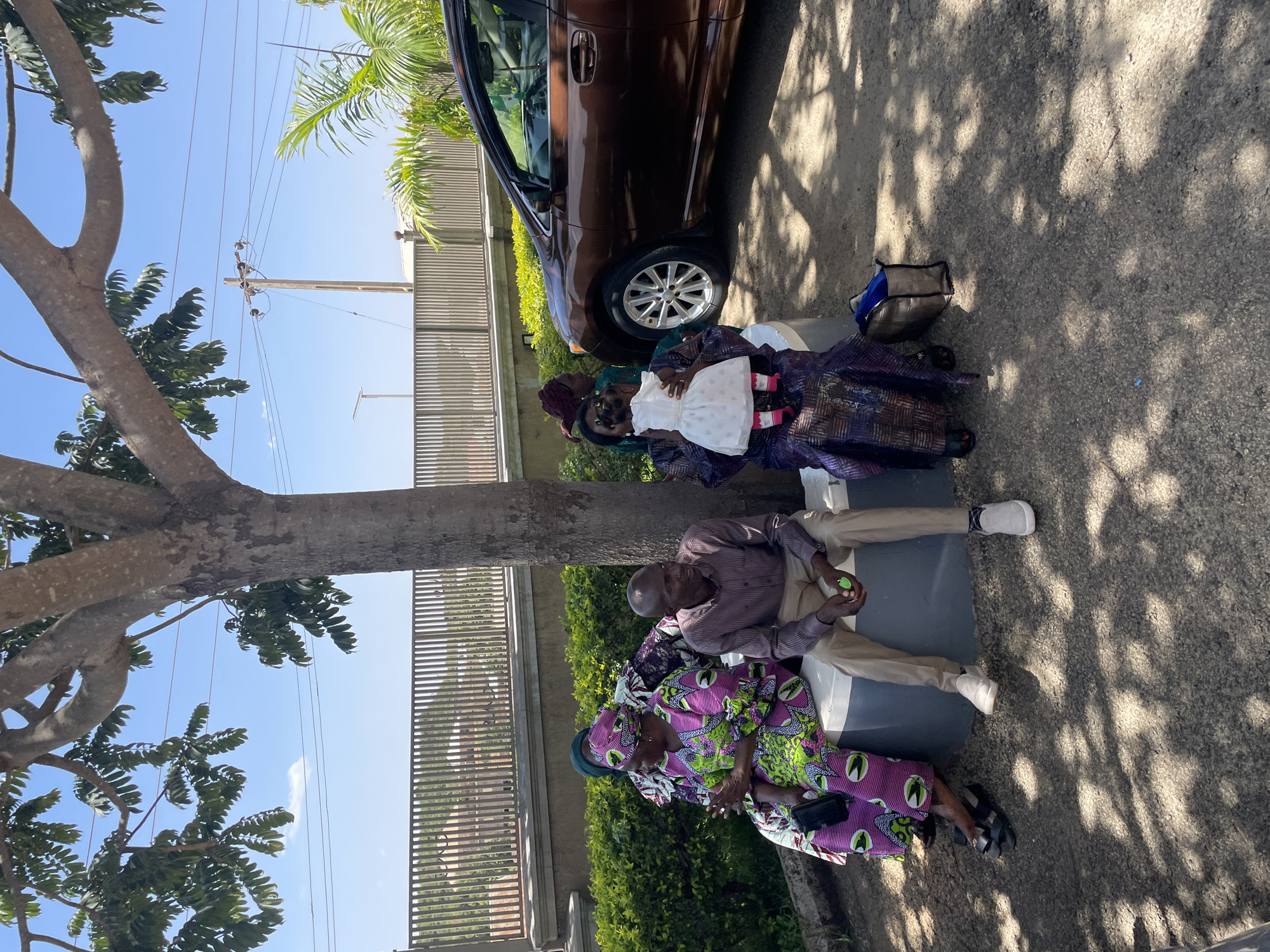 Four people sit on a concrete structure next to a brown car. One woman holds a baby dressed in white. One man is holding a small green object in his hand. A tree is nearby, casting shadows on the ground. The location is Jos, Nigeria.