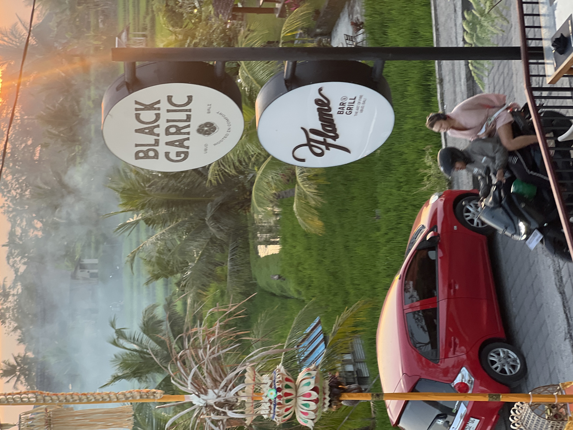 This is an eye-level, medium shot of a street scene in Tegallalang, Indonesia, at sunset. In the foreground, a red Honda Brio hatchback is parked on the left, its rear end facing the viewer. A black scooter carrying two people is to the right of the car, moving away from the viewer. The driver wears a helmet, and the passenger, a woman, is dressed in a pink robe. In the background, lush green rice paddies stretch into the distance, with palm trees and a few buildings interspersed. Smoke drifts across the scene, likely from a nearby fire or cooking. Two circular signs are mounted on a pole; the top one reads "BLACK GARLIC" with "Ubud, Bali" and a stylized garlic bulb beneath. The bottom sign reads "Flame" in script, with "BAR & GRILL" and "THE ART OF FIRE UBUD, BALI" below. A decorative Balinese umbrella-like object with colorful ornaments is positioned on the left side of the frame. The setting sun casts a warm glow over the scene, with rays of light visible in the upper right corner.