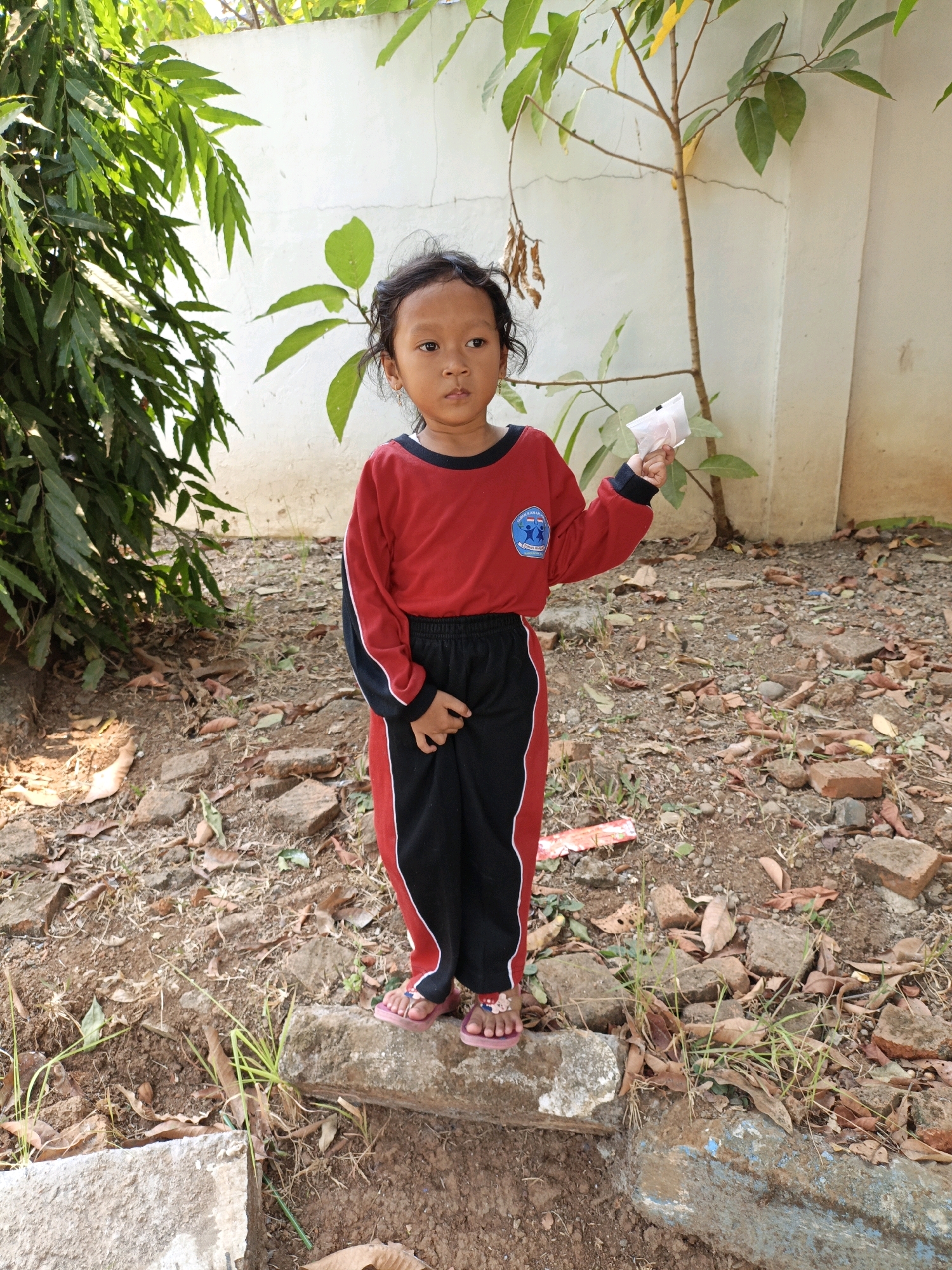 A young girl, appearing to be of Southeast Asian descent, stands outdoors in what seems to be a rural or semi-rural setting. She is wearing a red and black uniform with a blue and white emblem on the chest. Her hair is dark and curly, and she is holding a small, white object in her right hand. She is standing on a low, flat stone or concrete block, with a background of dry leaves, dirt, and scattered bricks. To her left, there is lush green foliage, and behind her, a light-colored wall and a slender tree with green leaves. The lighting suggests it is daytime, with sunlight casting shadows. The scene is consistent with a yard or an open area near a building in a warm climate, possibly in Kuwasen, Indonesia, given the vegetation and architectural style of the wall.