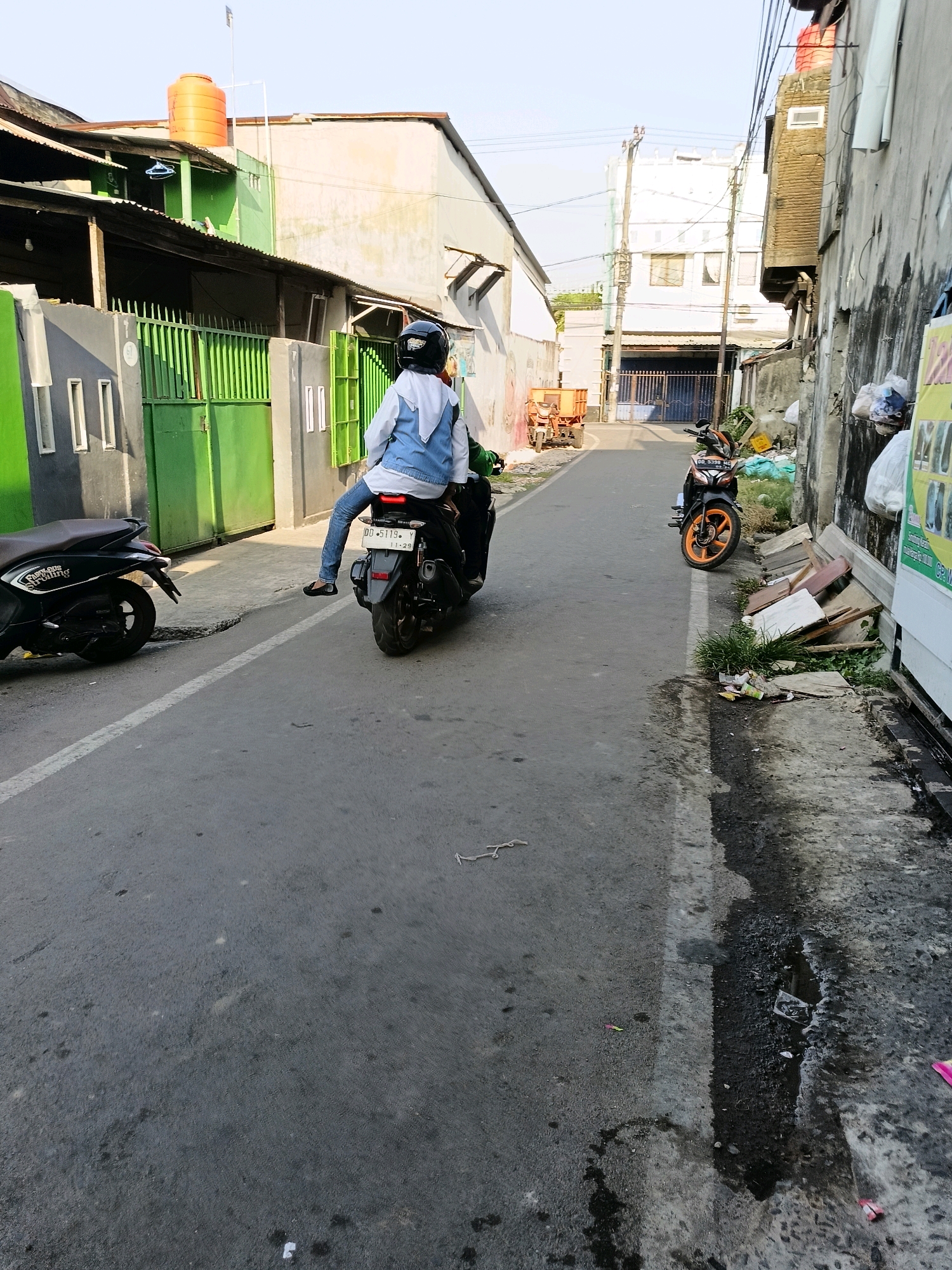 The image depicts a narrow street in an urban environment, likely within the city of Makassar, Indonesia, given the style of the buildings and the presence of motorcycles. In the foreground, a person wearing a blue jacket and jeans is riding a black motorcycle. They are also wearing a black helmet and a white hijab. To the left of this motorcycle, another black scooter is parked. The street itself is paved with asphalt and shows signs of wear and tear, with some water stains and debris visible. On the right side of the street, a wall made of concrete blocks runs alongside, with various items piled against it, including pieces of wood and what appears to be rubbish. In the background, buildings of varying heights and architectural styles are visible, some with corrugated metal roofs and others with more modern concrete facades. Power lines crisscross above the street, and a bright orange water tank is perched on top of one of the buildings. The overall impression is that of a lived-in, somewhat cluttered urban alleyway.