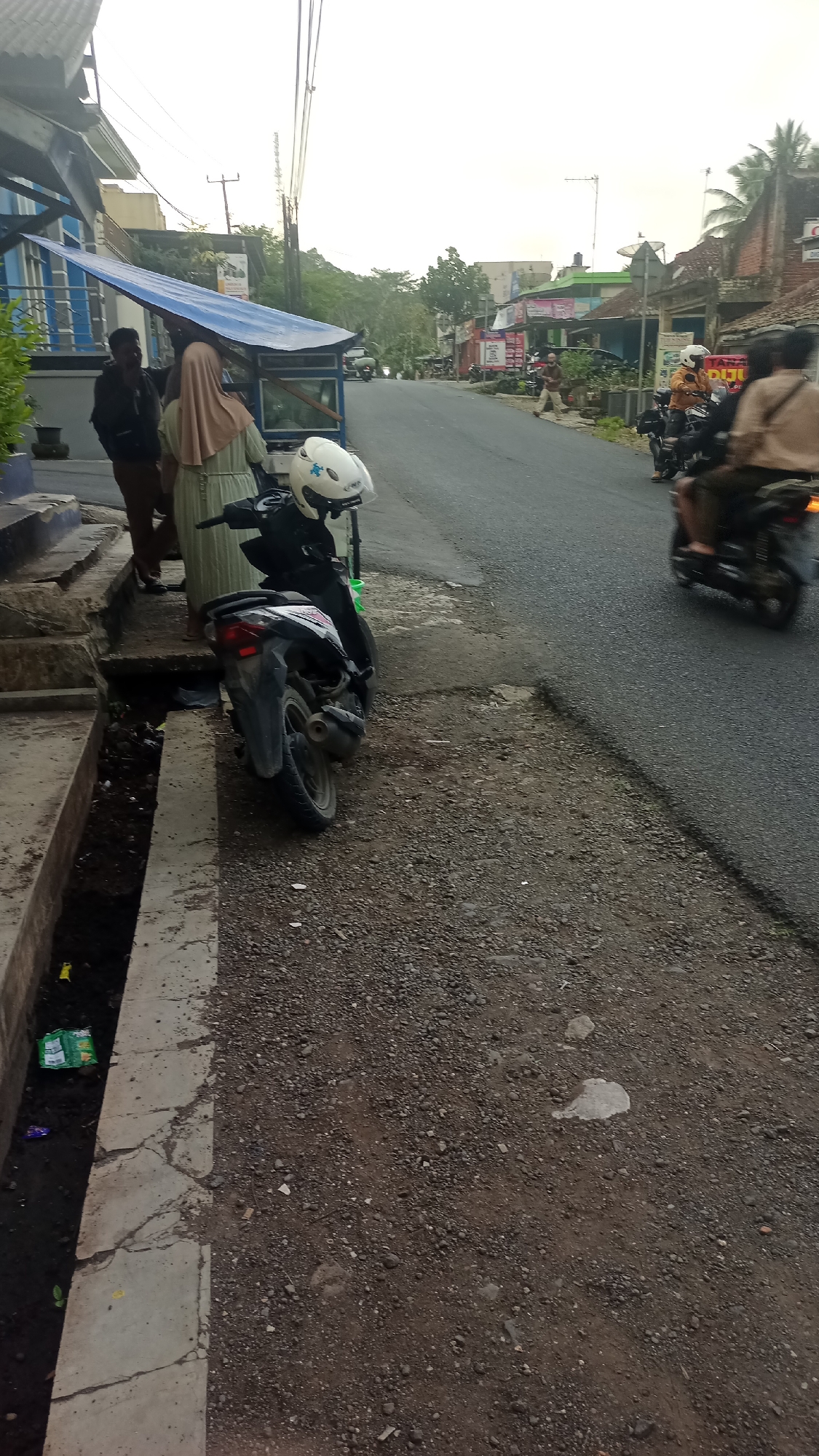 The image depicts a street scene in Karyamandala, Indonesia, likely during the late afternoon or early evening due to the soft lighting and overcast sky. On the left side of the frame, a blue awning covers a small structure, possibly a food stall or shop. Two individuals are standing near this structure; one is a man in dark clothing and the other is a woman wearing a light-colored headscarf and a patterned dress. A white helmet rests on the handlebars of a parked scooter in front of the awning.
The street itself is asphalt and slopes slightly upwards. Several motorcycles are visible on the road, with one rider in a brown jacket and helmet in the mid-ground and another rider in the background wearing a light brown jacket. The right side of the street is bordered by a gravelly area with scattered rocks and dirt. Buildings line the street on the right, some appearing to be shops or houses with various colored facades and signage, including what appears to be a sign for "DIJU." Trees and foliage are present along the sides of the street and in the background, indicating a semi-rural or suburban setting. A drain or ditch runs along the edge of the sidewalk on the left, containing some debris, including a green plastic bag and other small items. The overall atmosphere suggests a typical day in a local Indonesian community.