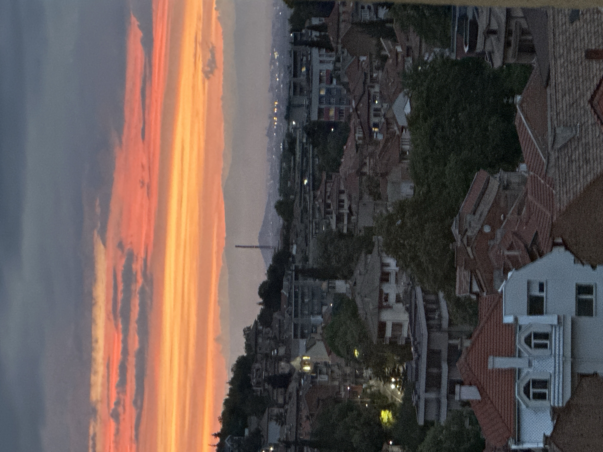 The image captures a city skyline at dusk, likely Asenovgrad, Bulgaria, characterized by a vibrant sunset illuminating the sky with hues of orange and red, contrasting with the darker, silhouetted cityscape below. The foreground features a cluster of residential buildings with distinctive red-tiled roofs, characteristic of Bulgarian architecture. Several structures display illuminated windows, suggesting evening activity within. In the mid-ground, more densely packed buildings, some appearing to be older, rise up a hillside. Trees dot the landscape, interspersed among the buildings. In the distance, a hazy mountain range is visible, with a tall, slender industrial chimney or tower standing out against the fading light. The overall impression is that of a typical urban settlement transitioning from day to night, with the natural beauty of the sunset providing a dramatic backdrop.